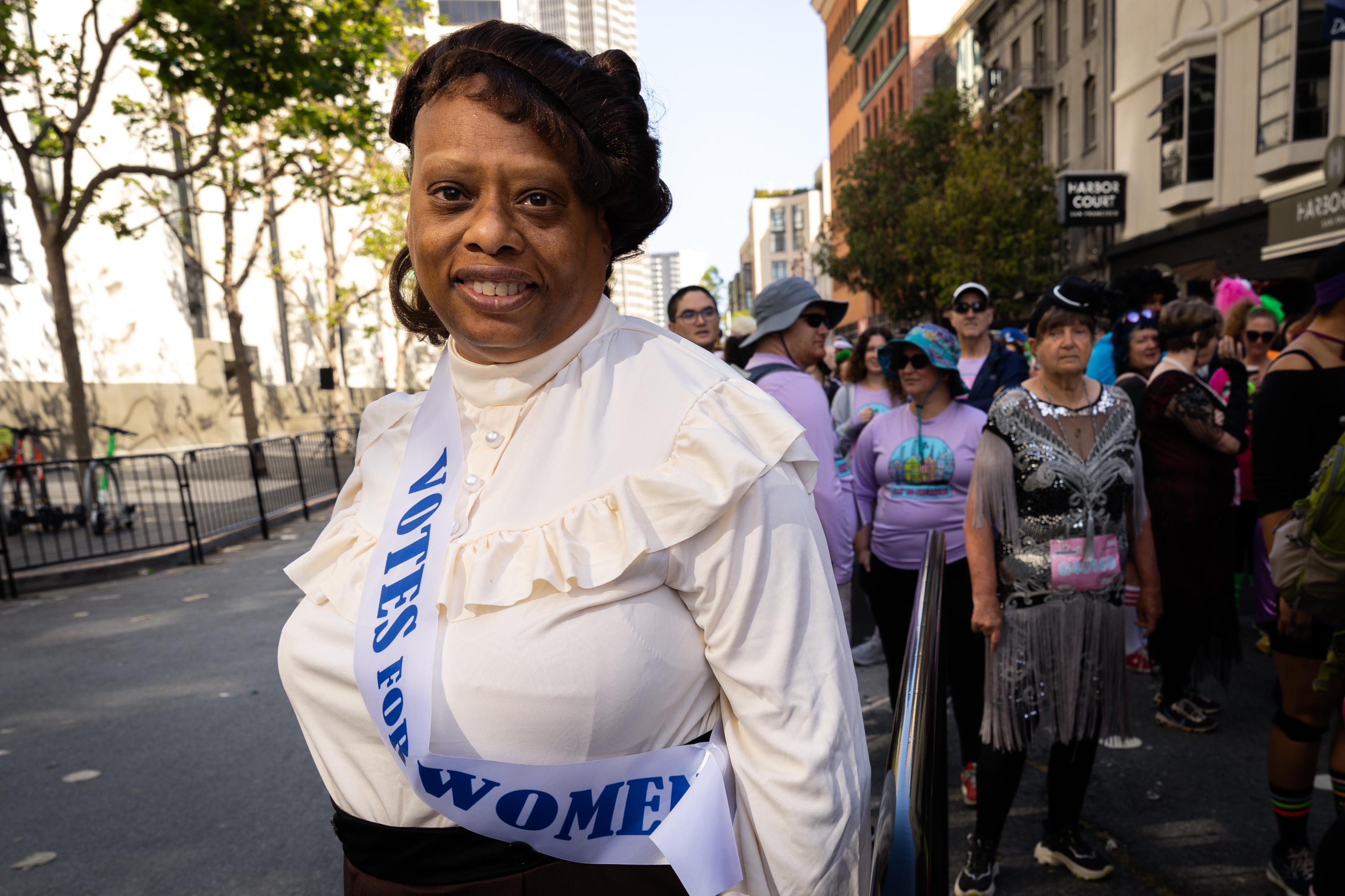 A woman in a historical white blouse and brown skirt stands in a crowd, wearing a sash that reads 'Votes for Women', smiling at the camera.