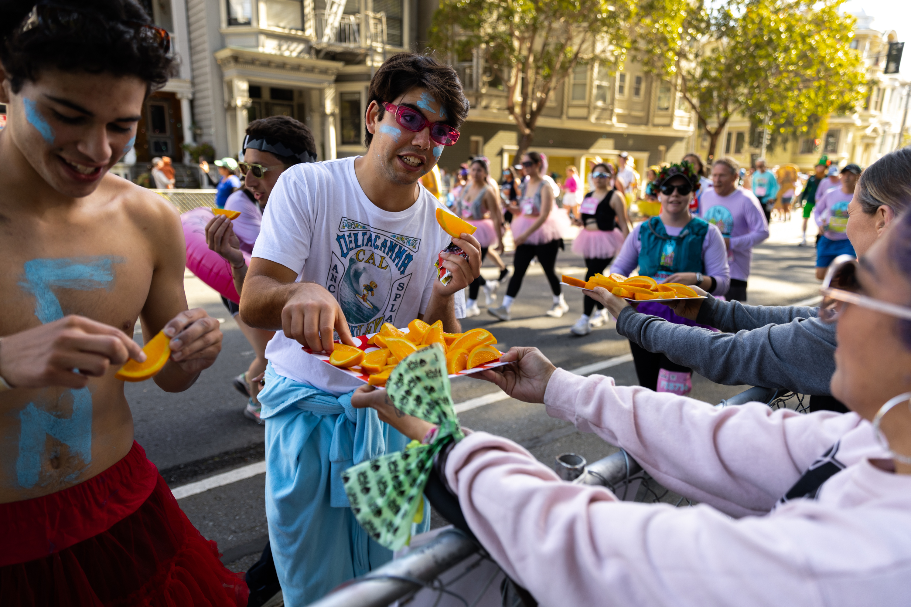 Participants grab orange slices from volunteers during a lively street event, sporting colorful attire and fun makeup.