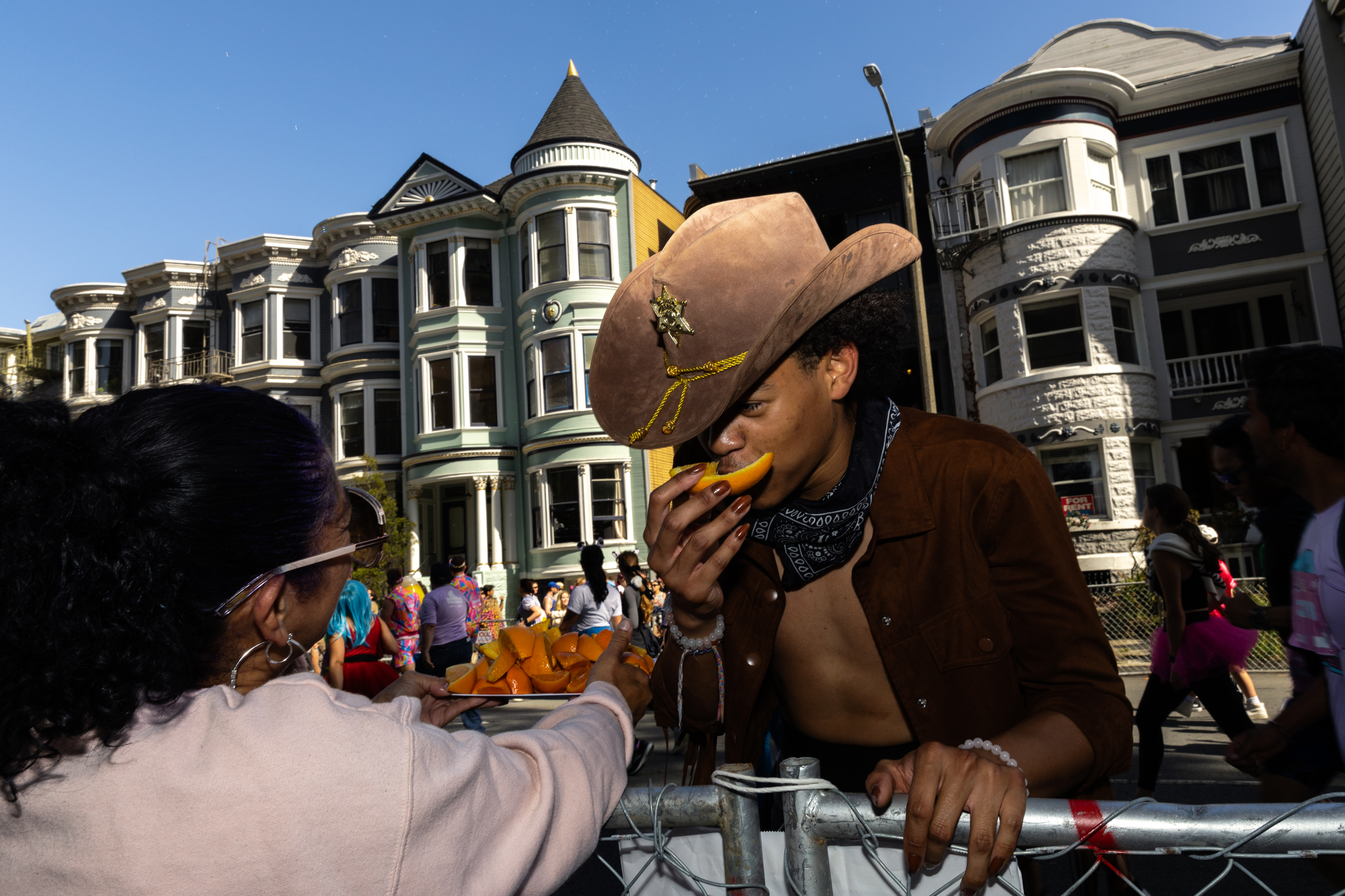 A person in a cowboy hat is eating an orange slice from a tray held by another. They are in front of colorful Victorian-style houses under a clear sky.