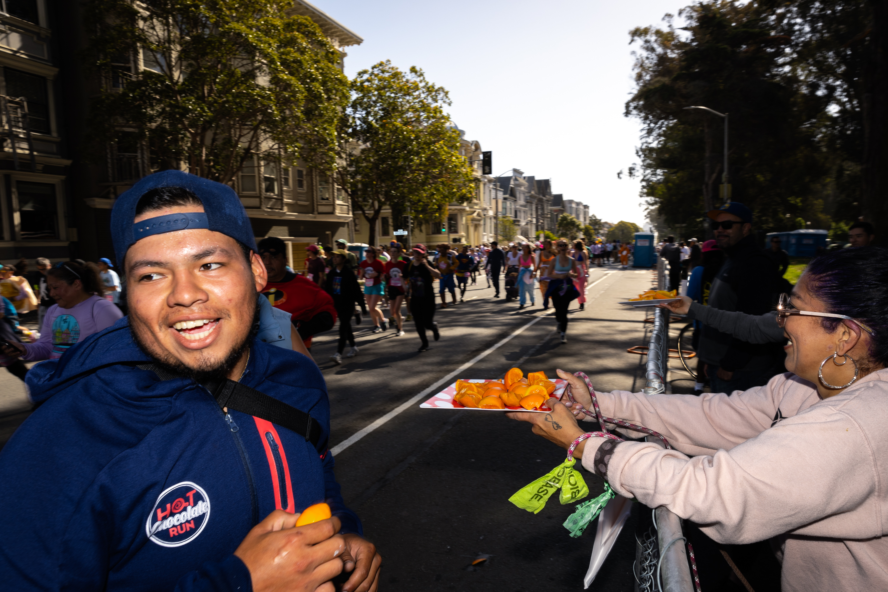 A smiling man in a blue cap receives orange slices from a woman at a lively street marathon lined with spectators and participants.