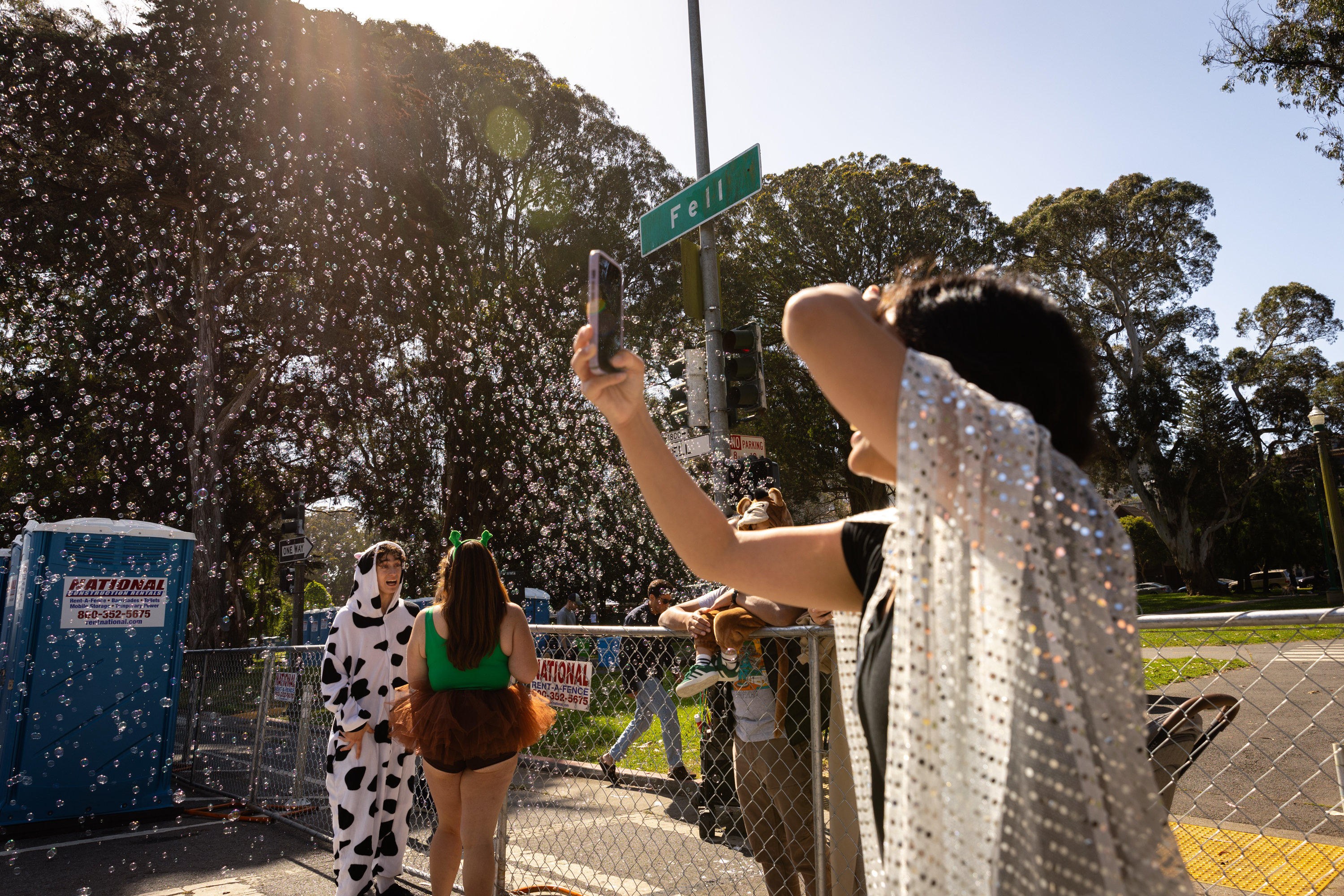 A crowd enjoys a sunny outdoor event, recording a flurry of bubbles in the air. People in playful costumes add festivity to the scene under a street sign labeled &quot;Fell.&quot;