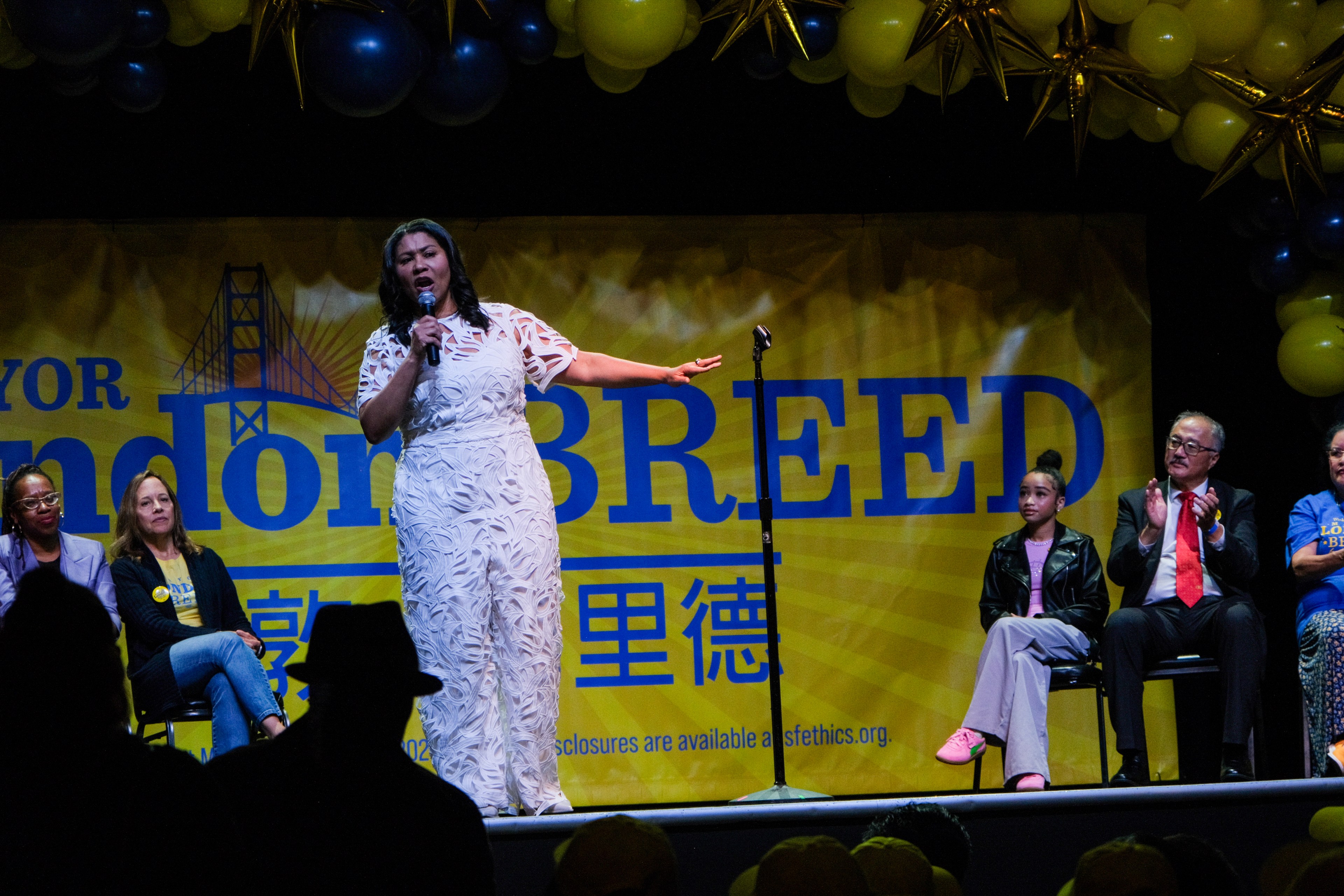 A woman in a white dress is speaking into a microphone on a stage with a yellow banner that reads "London Breed." Several people are seated behind her.