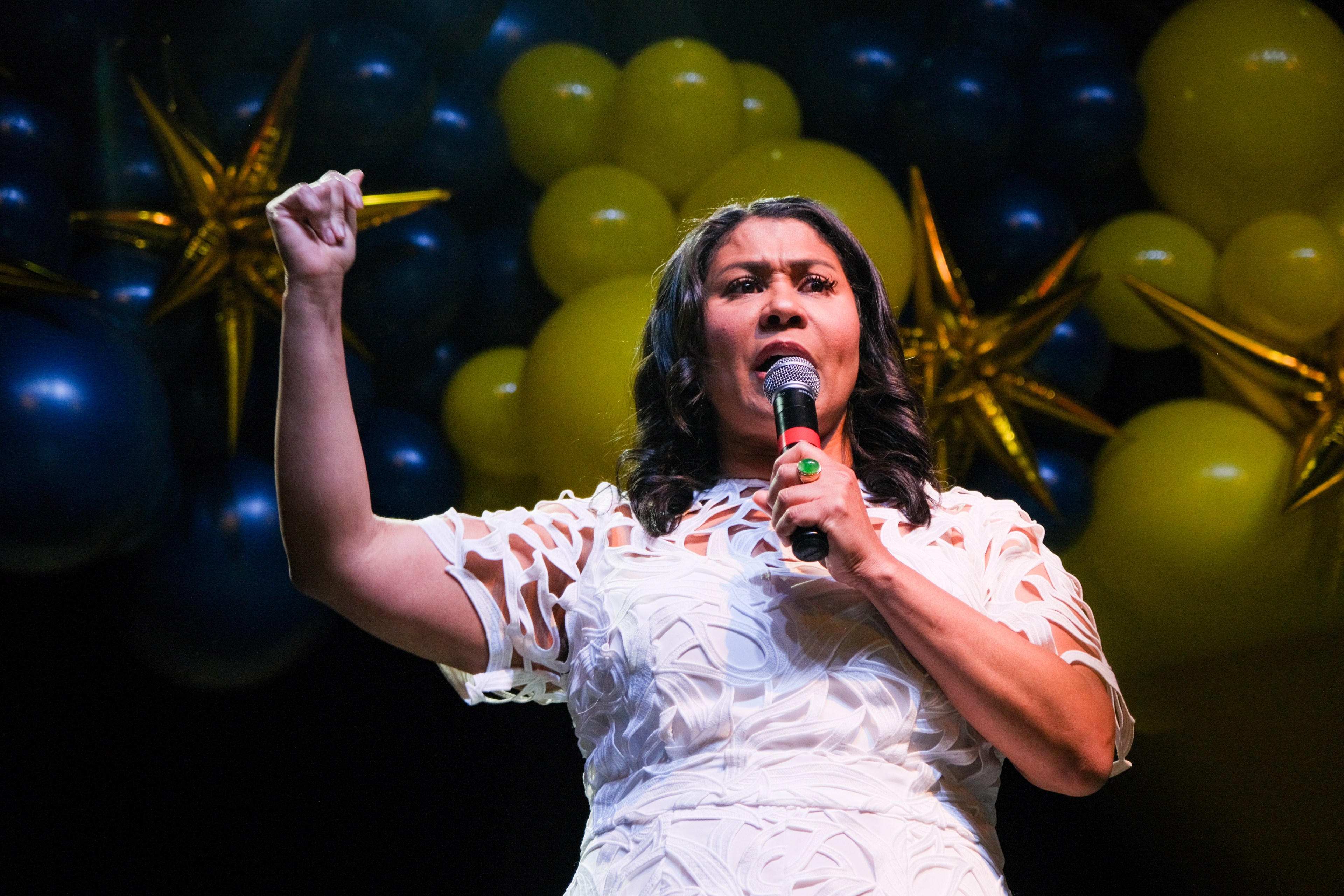 Mayor London Breed in a white patterned dress jumper into a microphone, passionately raising her fist, with festive gold and blue balloons in the background.