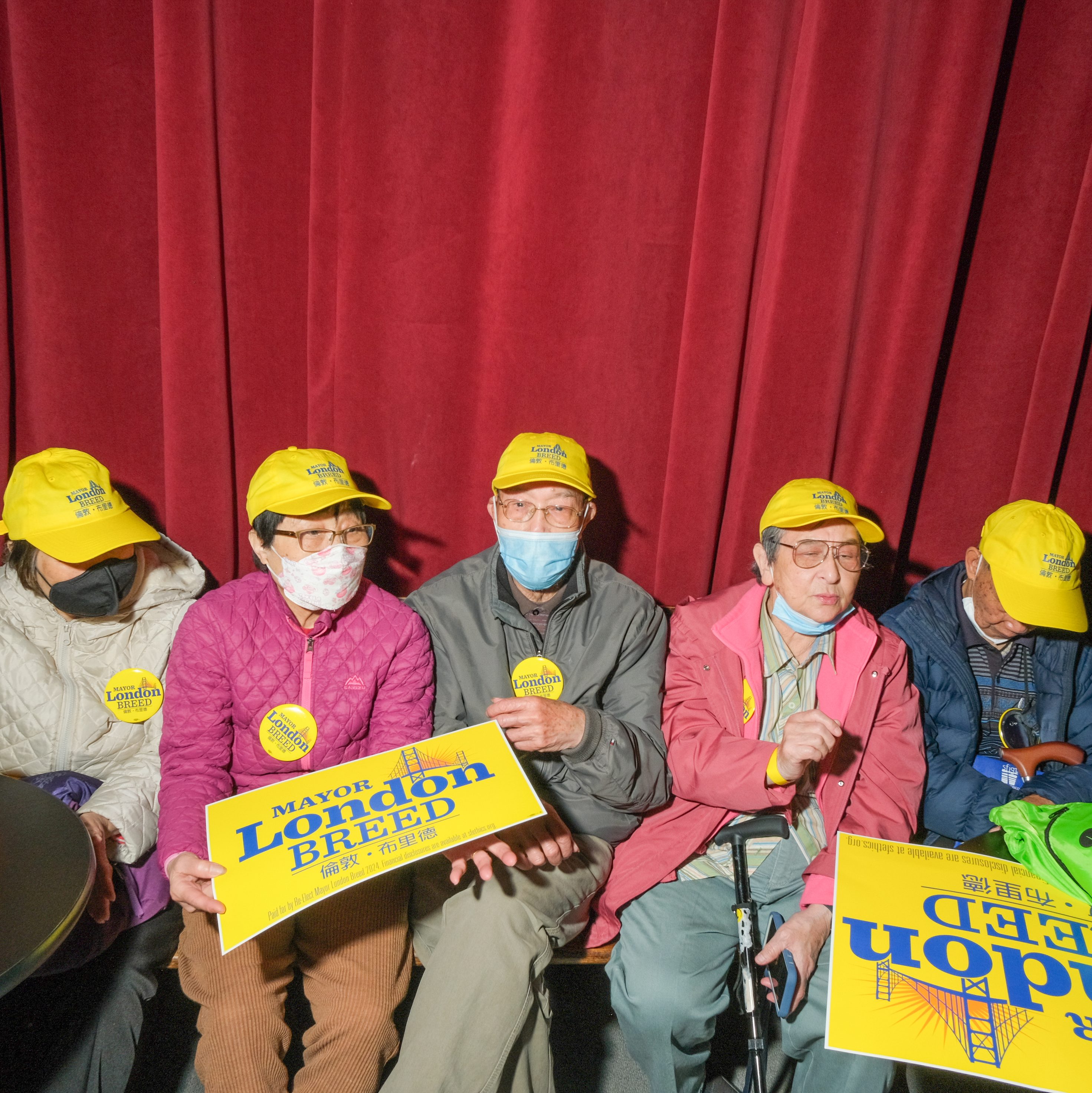A group of elderly people, wearing yellow caps and jackets, sit holding &quot;Mayor London Breed&quot; campaign signs by a red curtain. Some wear masks.