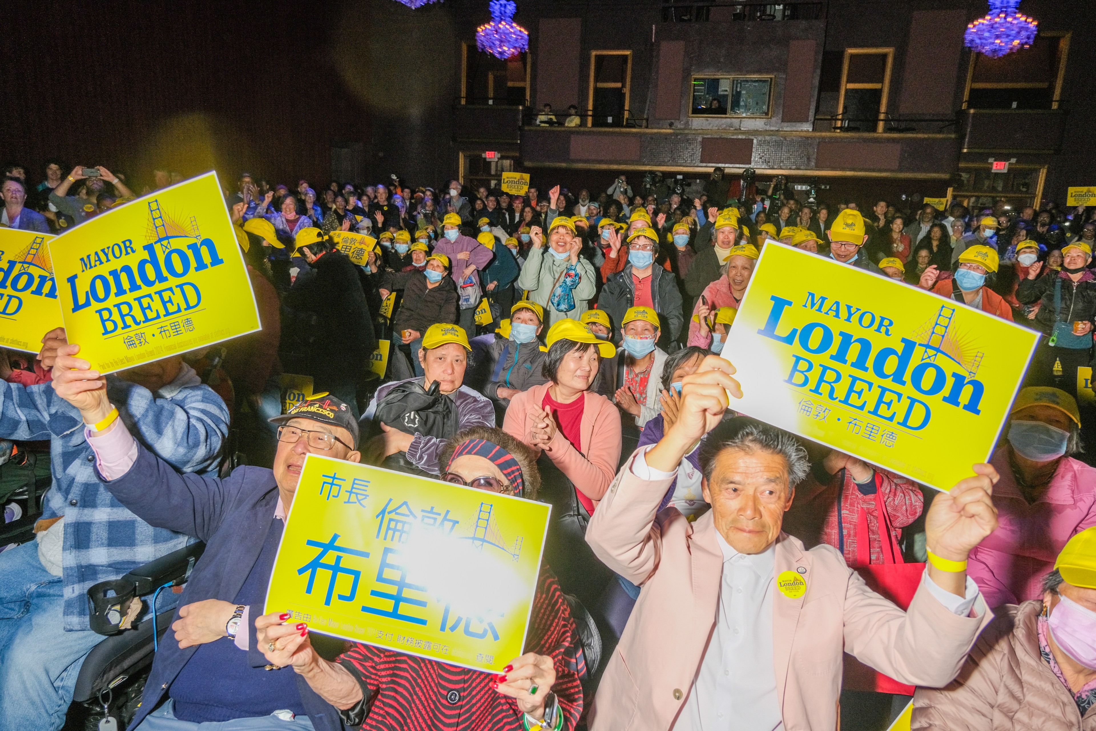 A crowd of joyful people, many wearing yellow hats, holds up signs supporting Mayor London Breed, in a well-lit indoor setting.