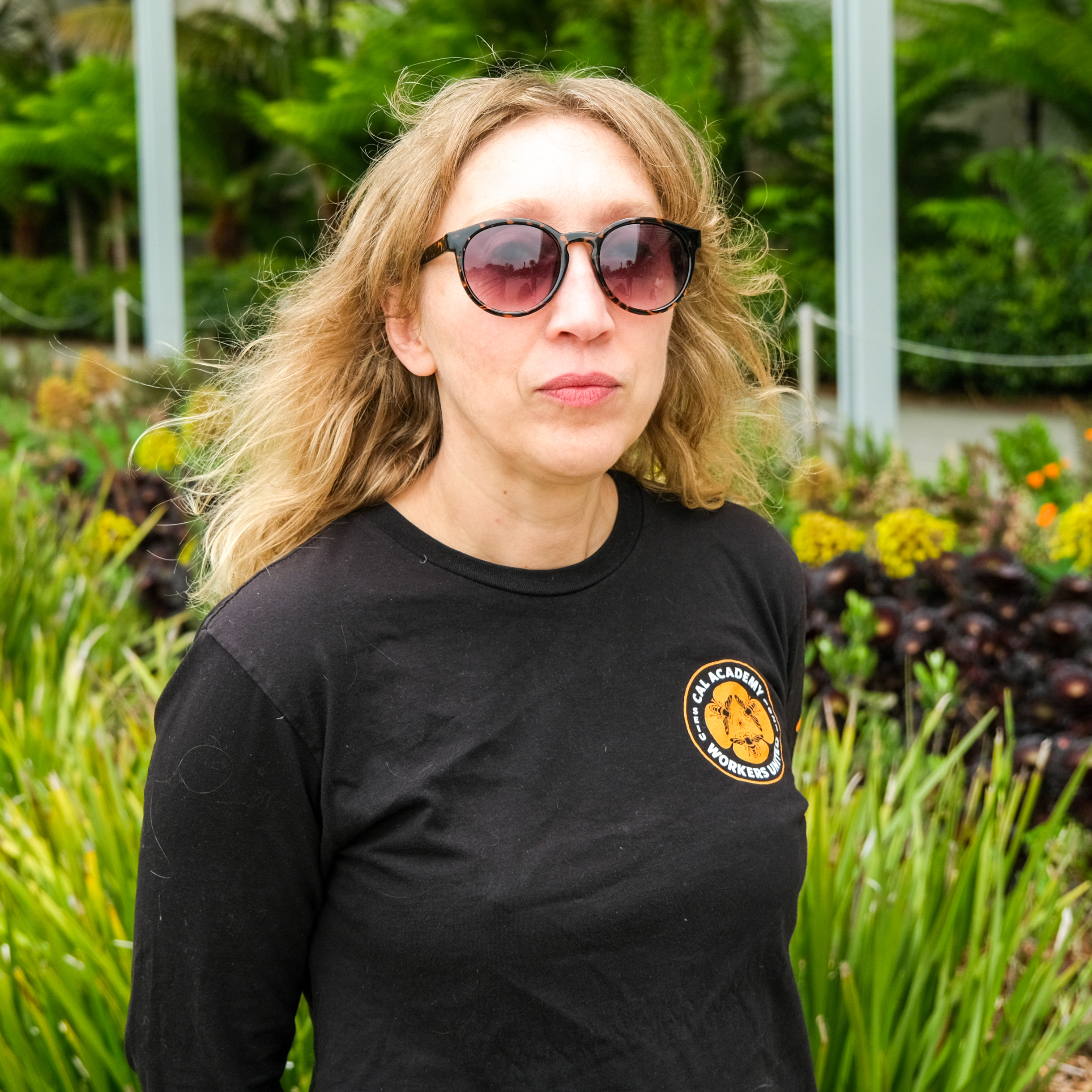 A woman in sunglasses and a black t-shirt stands in front of lush greenery, slightly glancing away from the camera.