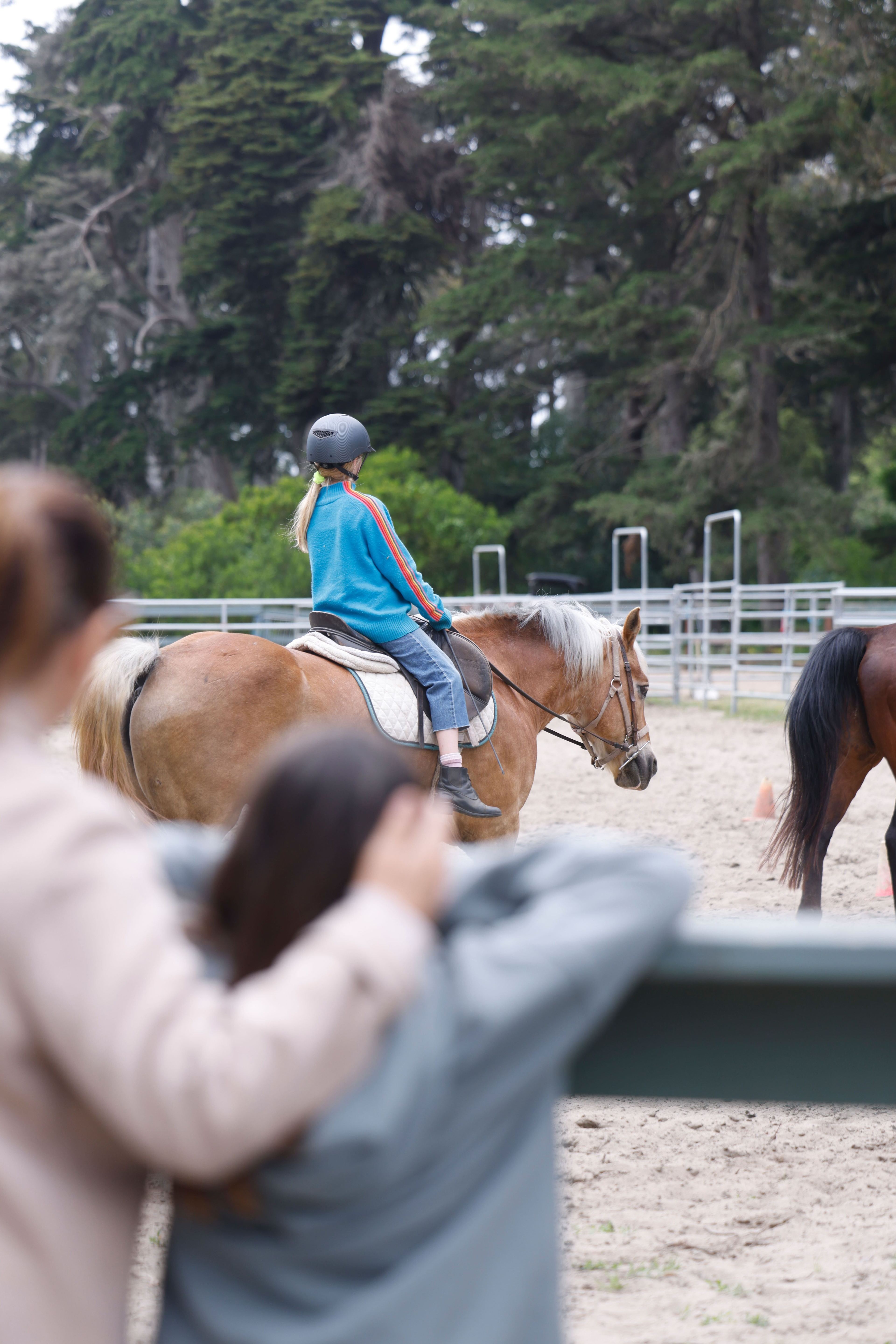 A person wearing a helmet rides a horse in a sandy area, while another person in the foreground watches. The background shows trees and a fenced area.