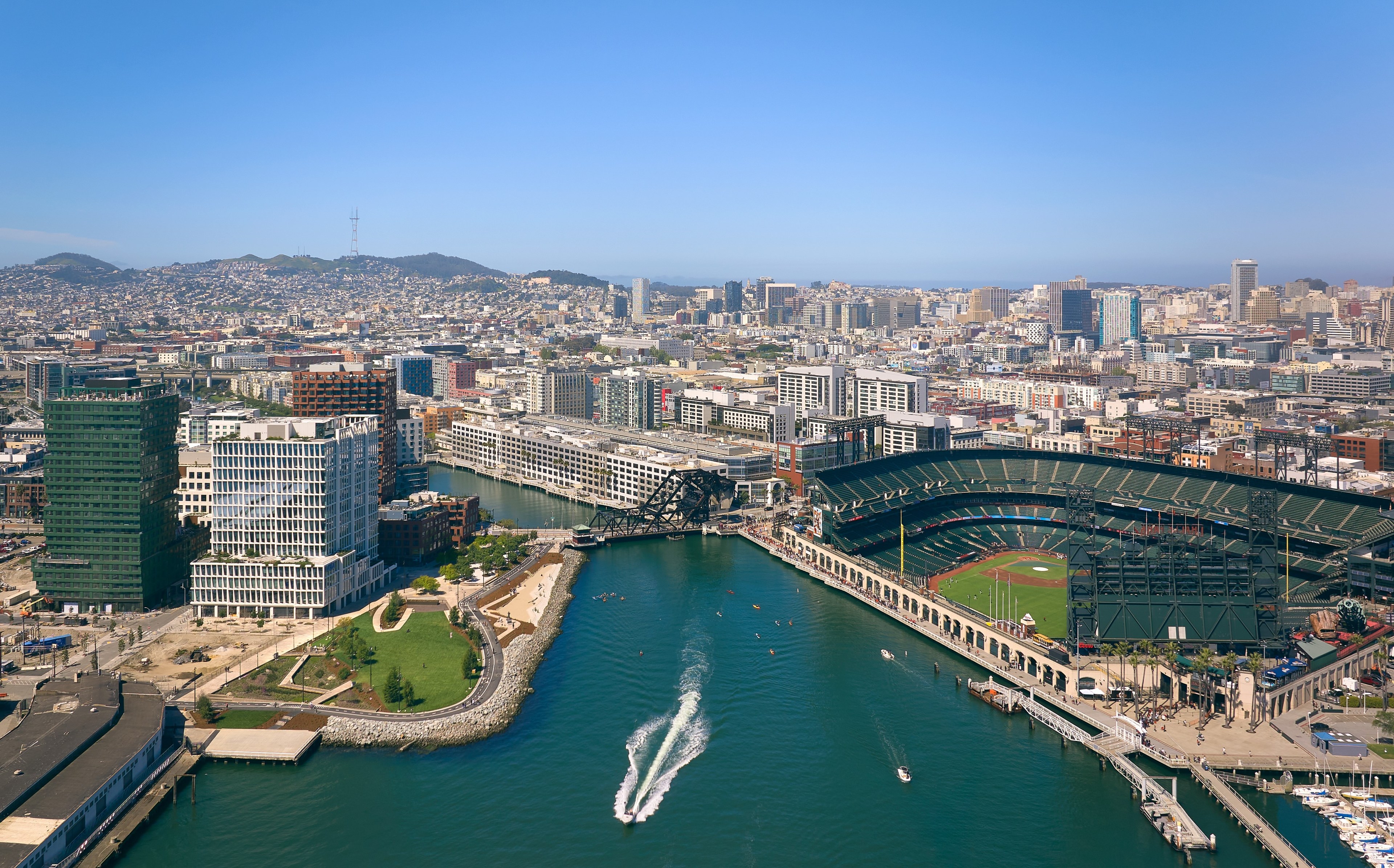 An aerial view of a city waterfront with a stadium, boats, modern buildings, and hills in the background.