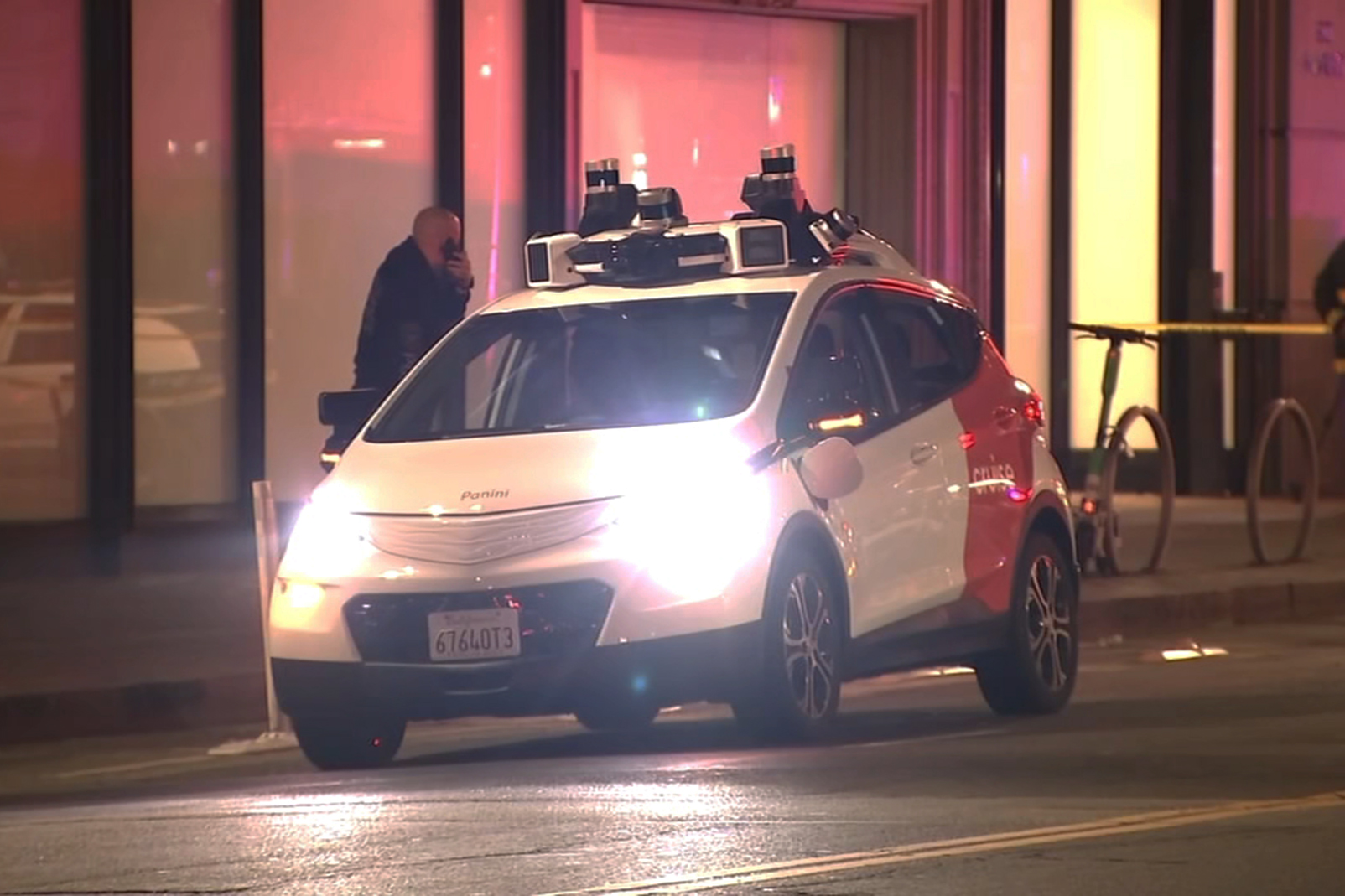 A self-driving car, equipped with multiple sensors on top, is parked on a city street at night, with a man standing nearby talking on a phone.