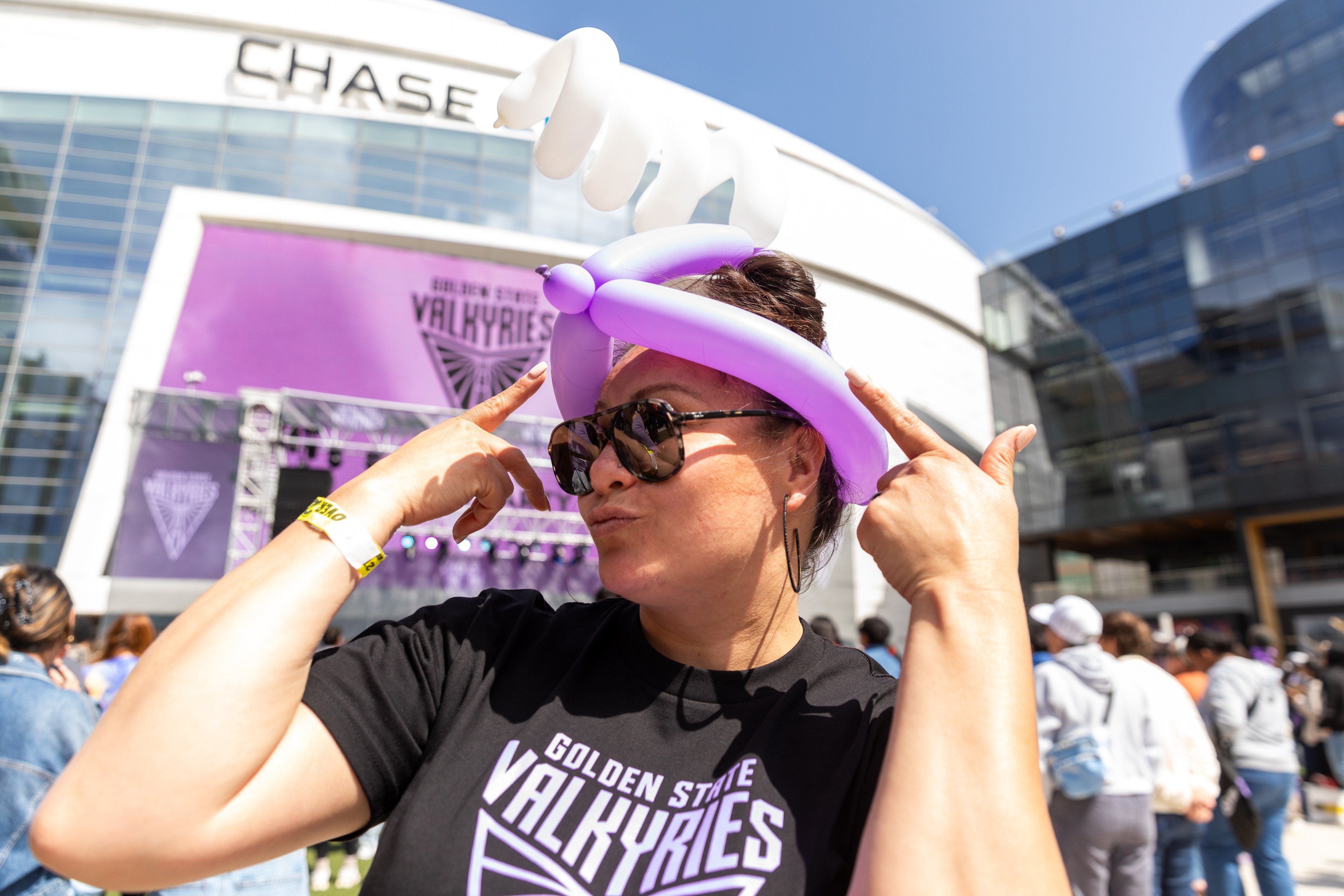 A woman gestures a peace sign, wearing sunglasses and a purple balloon hat at an outdoor event named "Golden State Valkyries" near the Chase building.