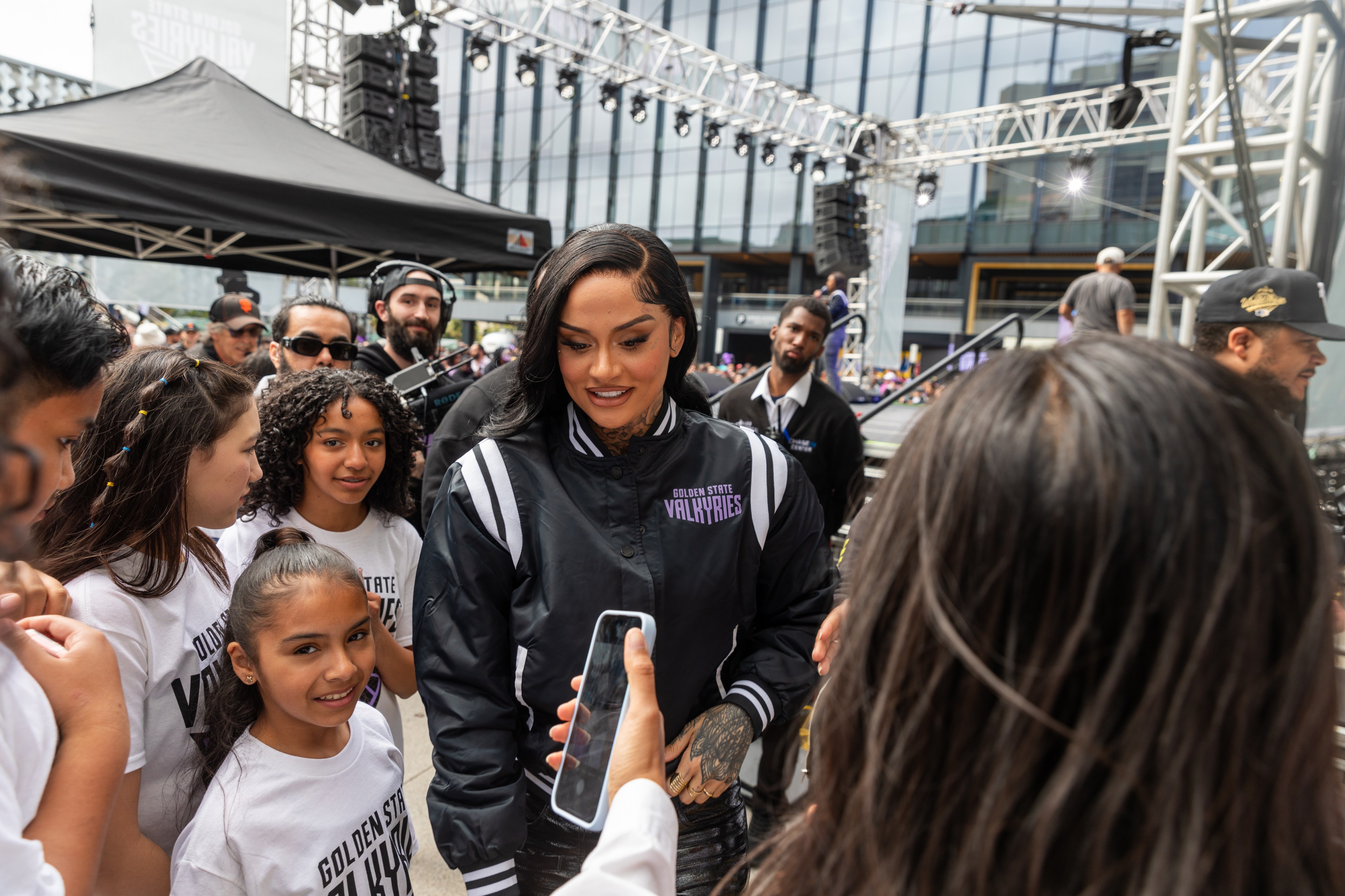 A woman in a sports jacket interacts with children in a crowded event, smiling as she looks at a phone held by a young girl.
