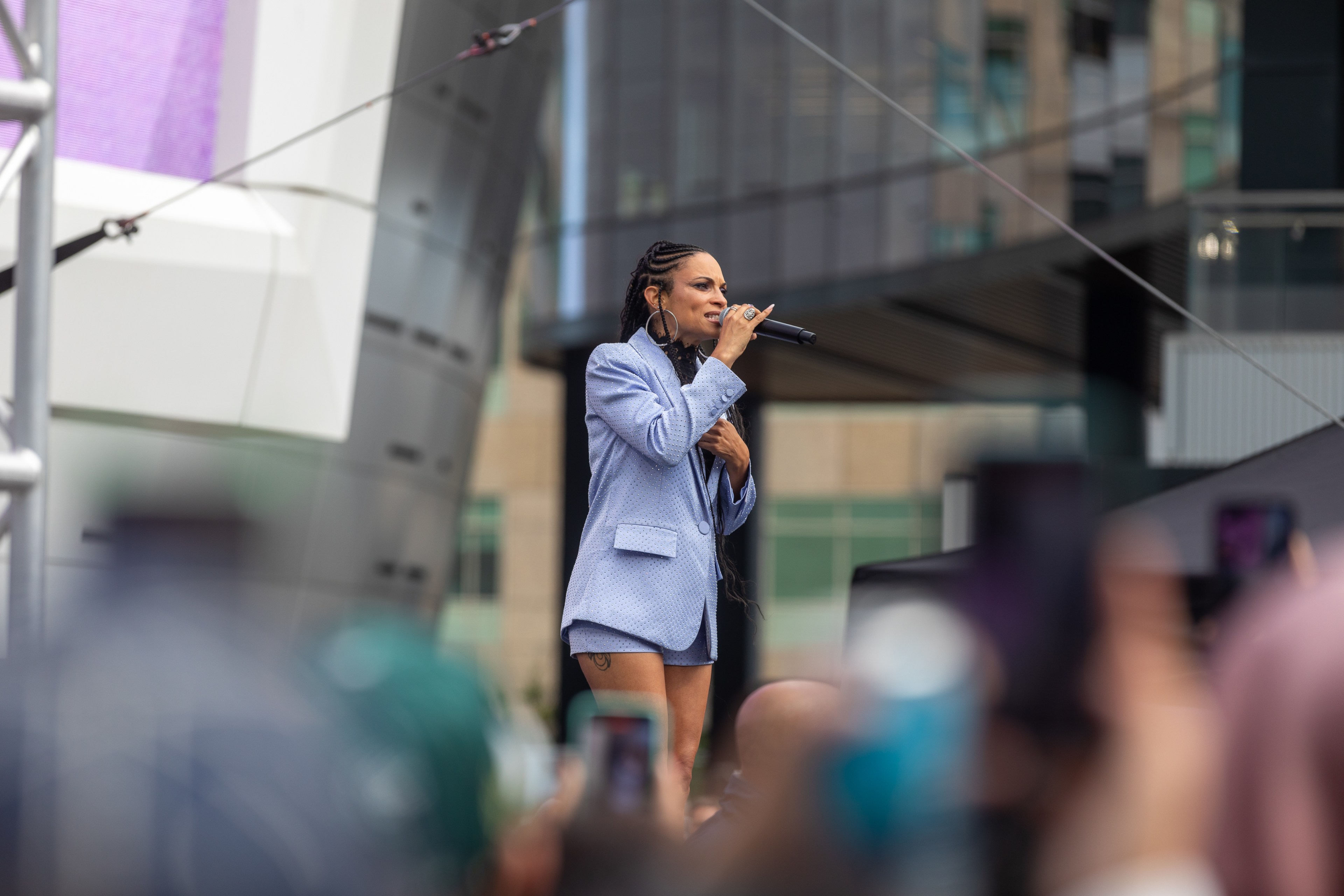 A woman is performing on stage in a city, holding a microphone and wearing a blue blazer, with an audience capturing the moment on their phones.