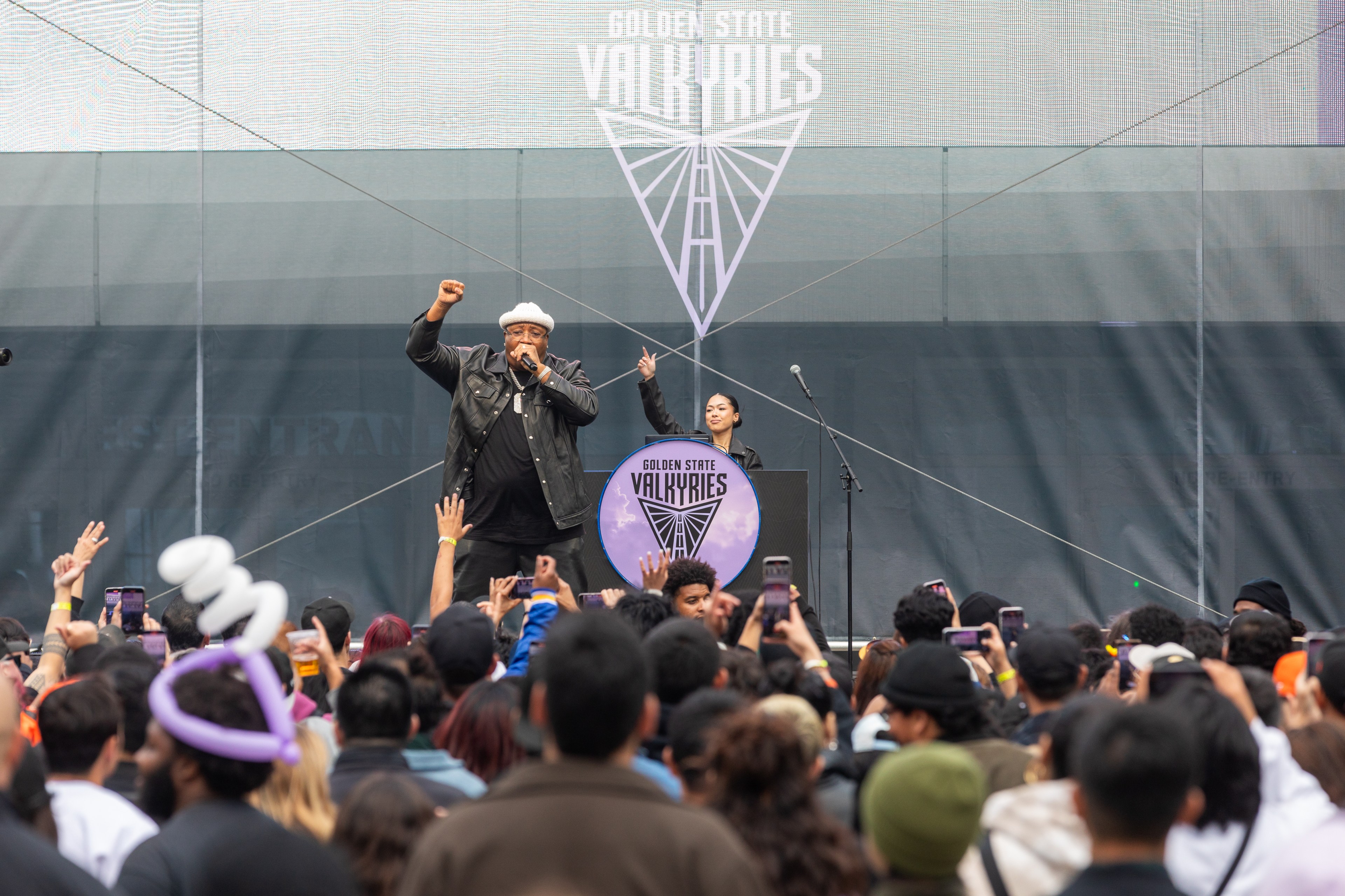 A performer in a black jacket and hat raises a fist on stage in front of a crowd, with &quot;Golden State Valkyries&quot; displayed on a large screen behind.