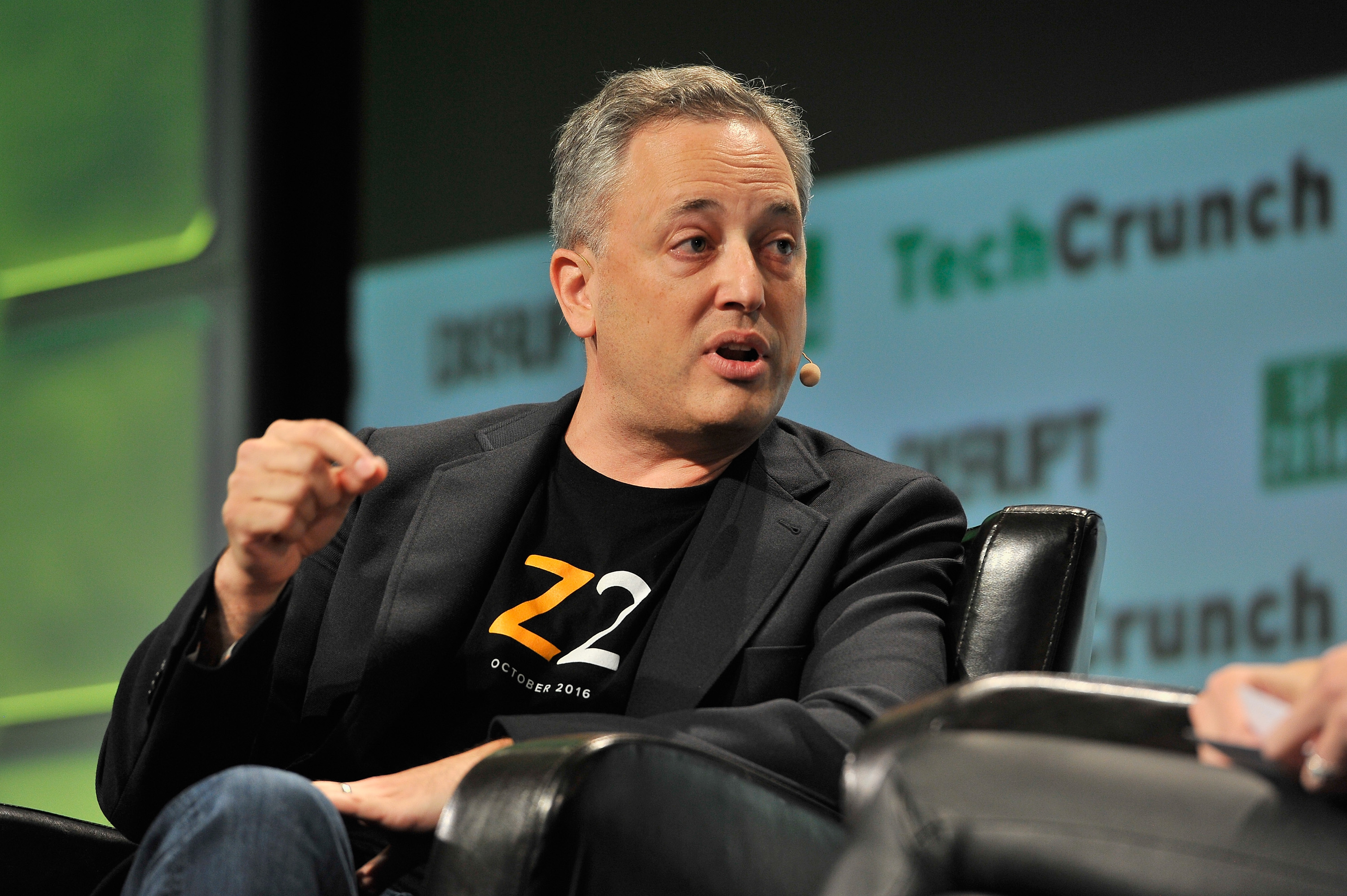 A man in a black blazer with a microphone headset is talking passionately on stage with TechCrunch logos in the background.