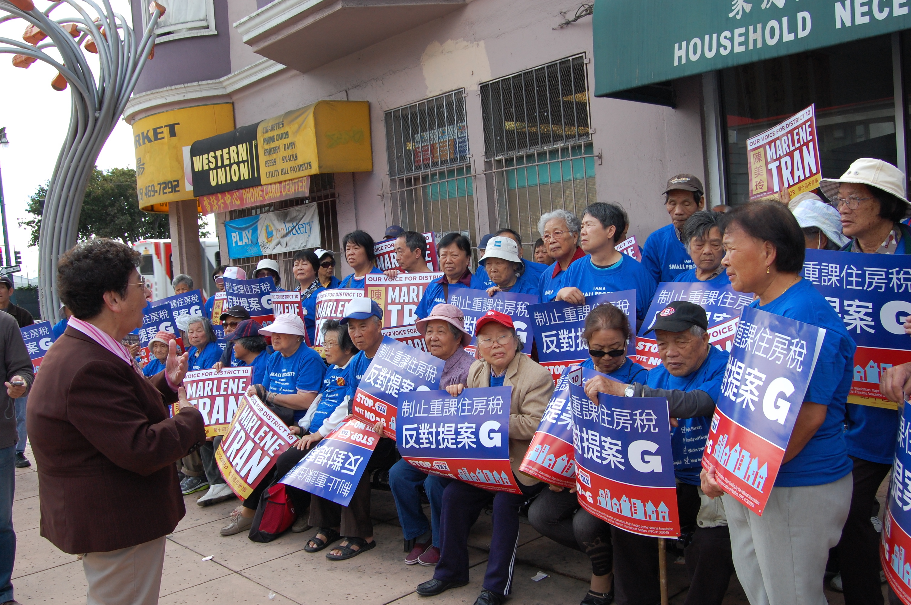 A group of elderly Asian people, holding political campaign signs, listen attentively to a speaker in an urban setting.