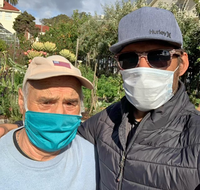 Two men, one in a cap and mask, another in sunglasses and mask, stand side by side outdoors.