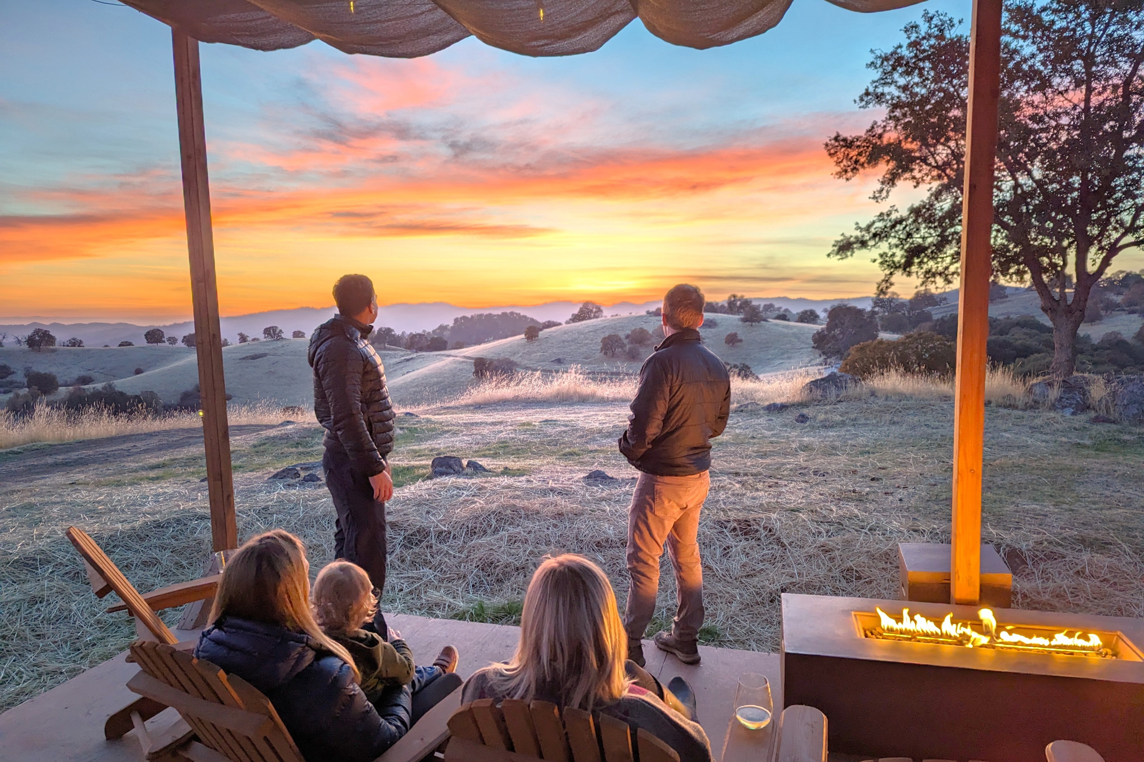 Five people under a canopy enjoy a scenic sunset view over rolling hills. Two stand looking out, while three sit by a modern outdoor fireplace, with drinks.