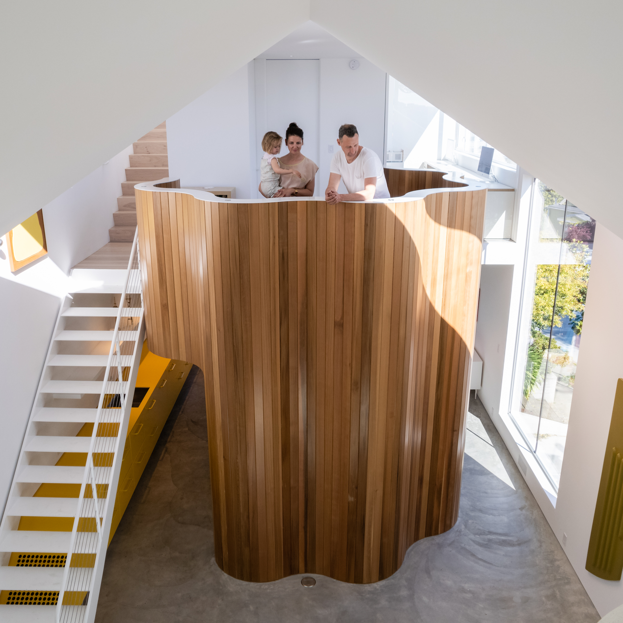 Three people converse by a unique curved wooden wall inside a modern, bright building with white stairs and triangular openings.