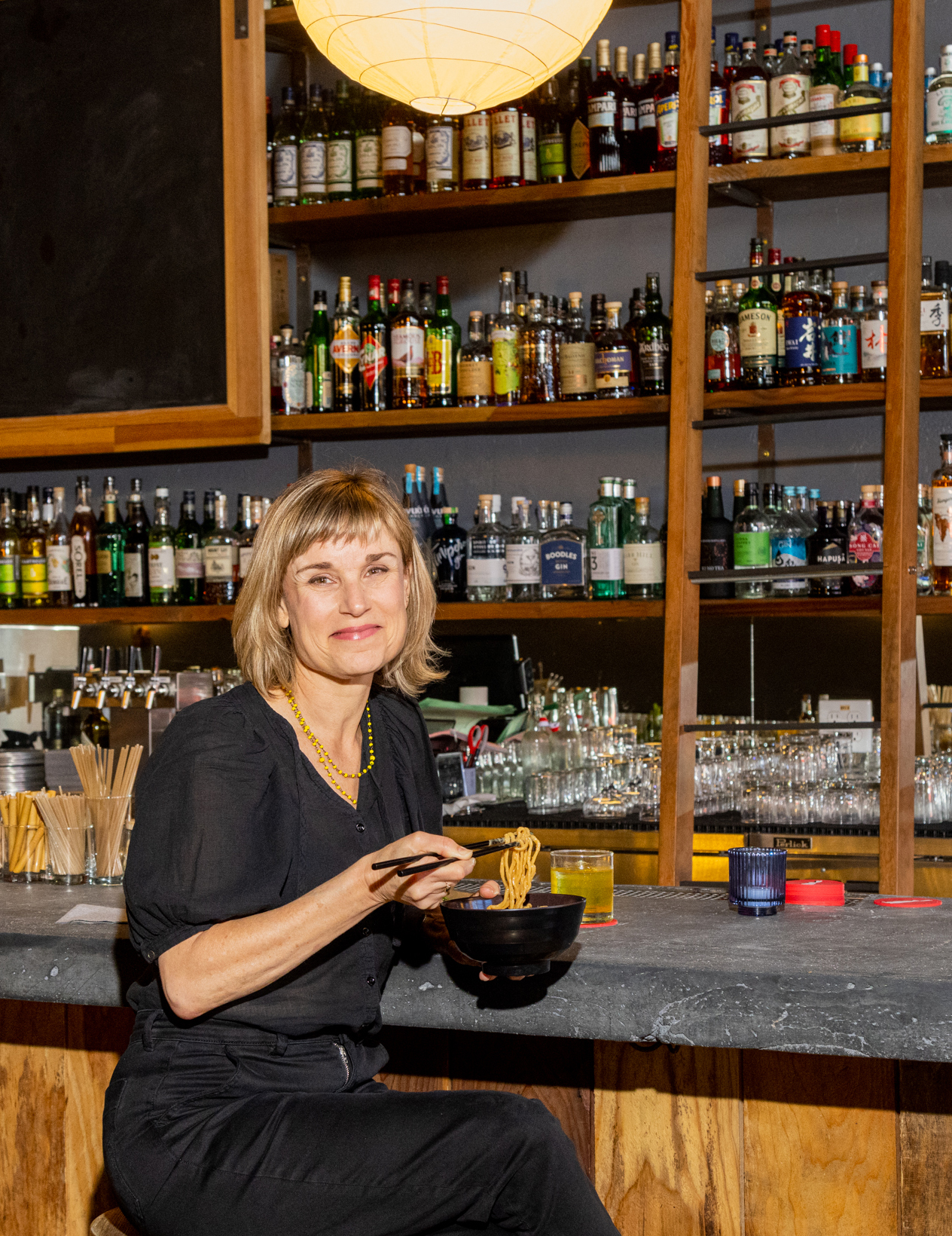 A woman is smiling, sitting at a bar with a bowl and chopsticks, shelves of liquor bottles behind her.