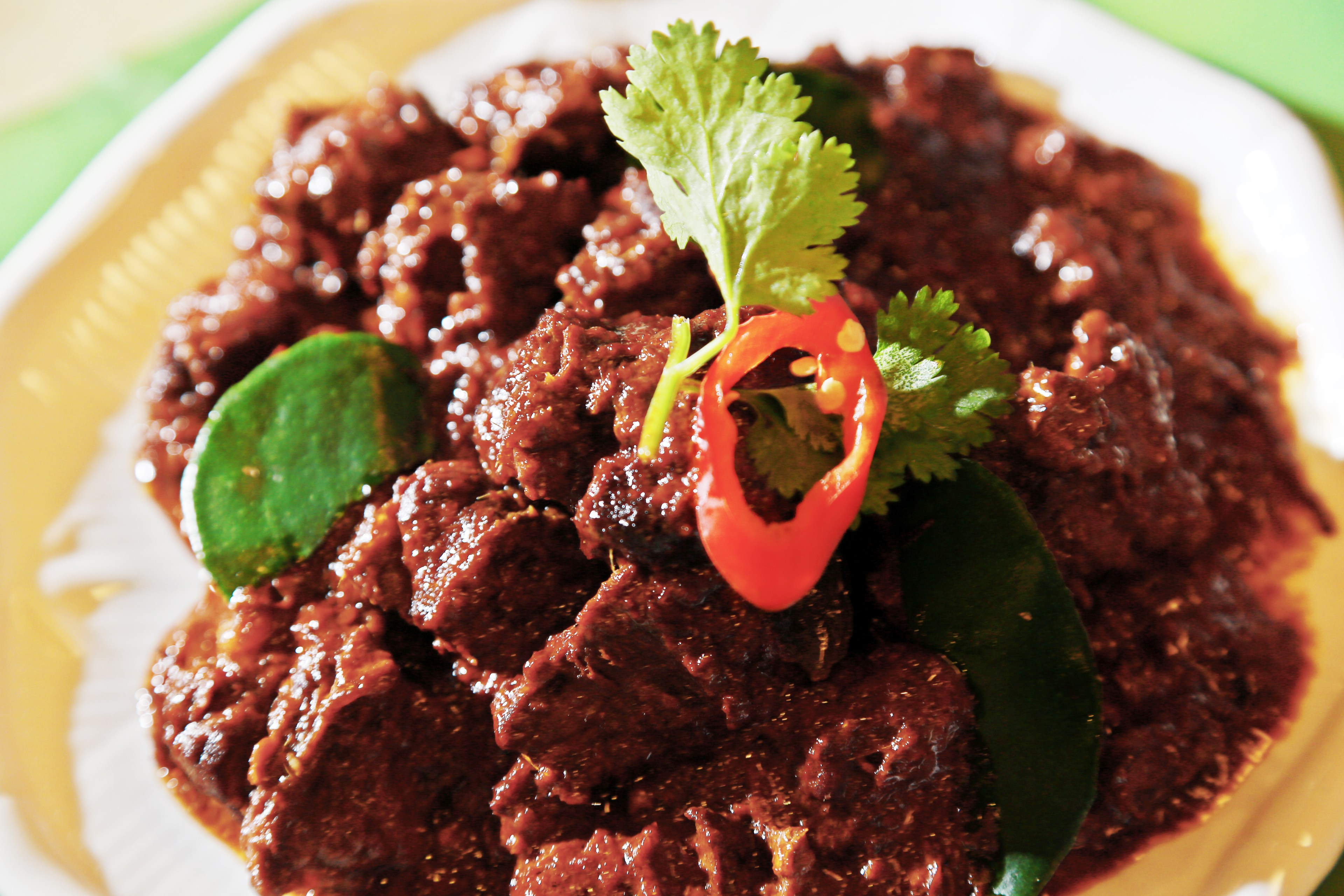 A dish with rich, dark rendang curry, decorated with fresh green lime leaves and a bright red chili slice, served on a yellow plate.