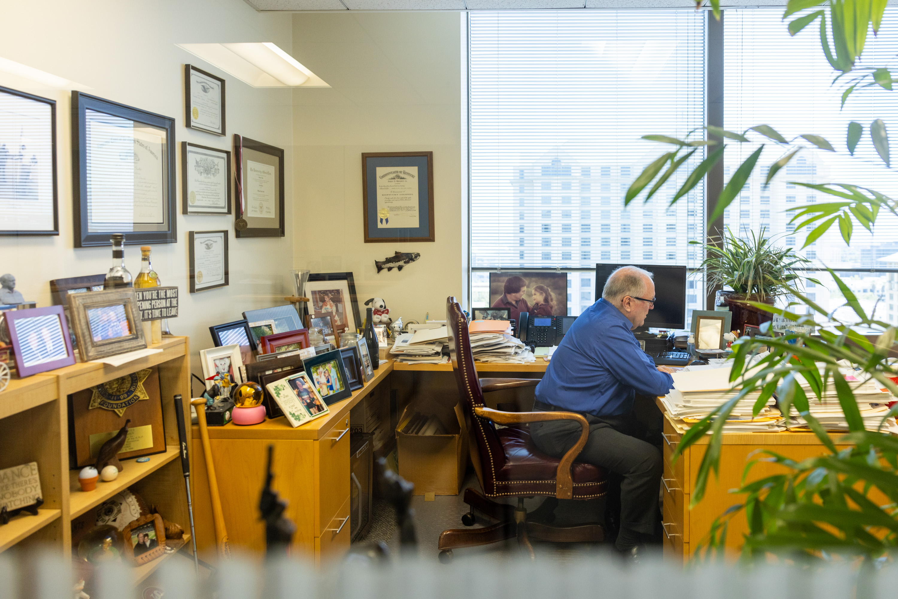 A man in a blue shirt works at a cluttered desk in an office. The room is filled with framed certificates, photos, and plants, with a city view visible through the windows.