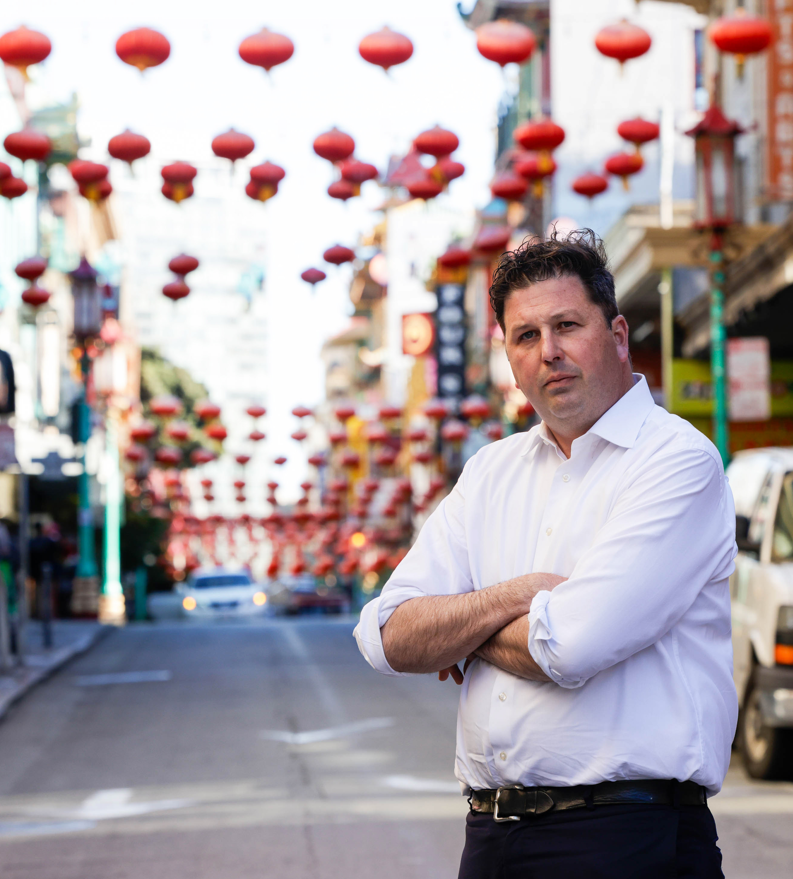 A man stands with arms crossed on a street lined with red lanterns above.