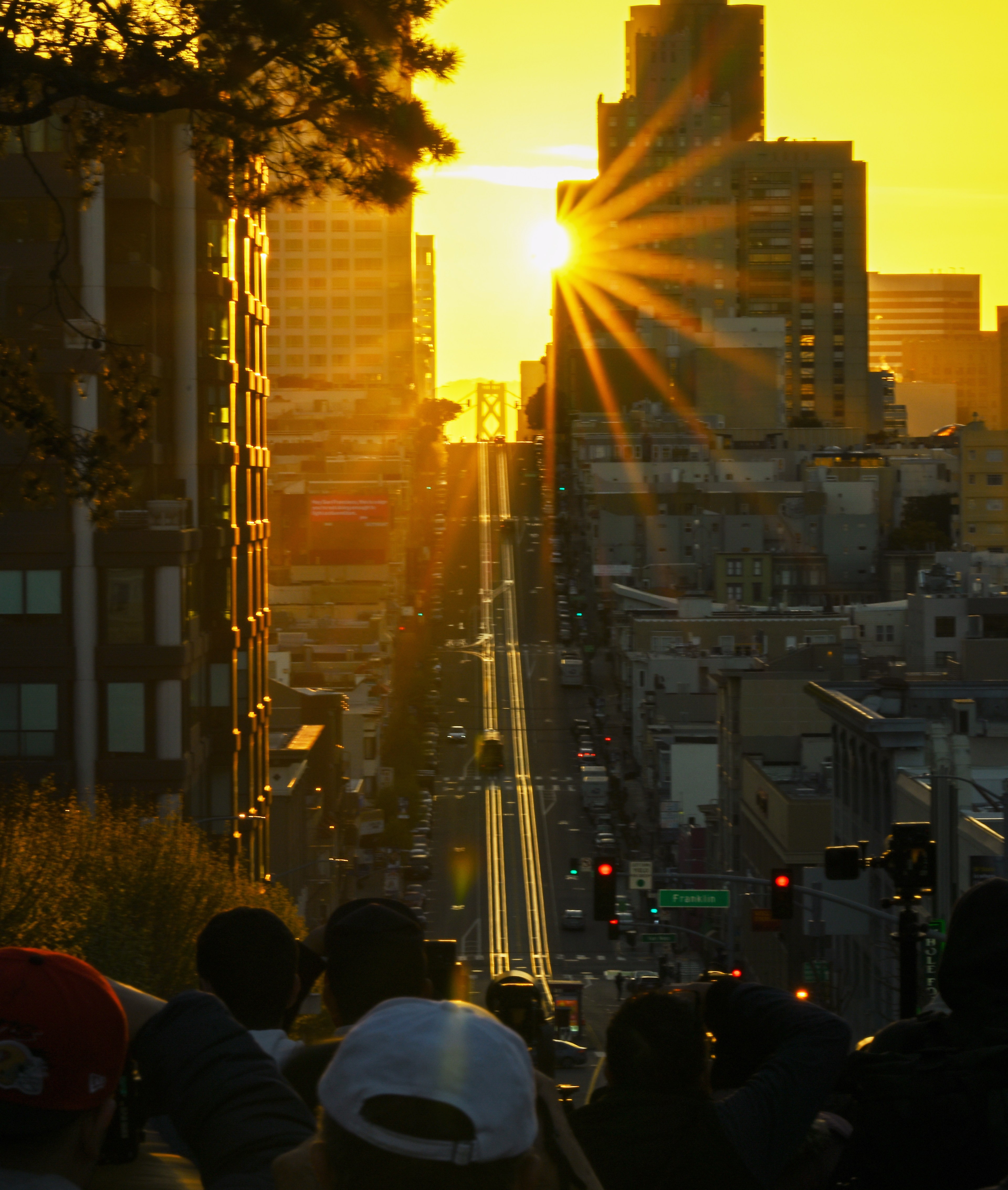 Sunset view down a steep city street, with the sun casting starburst rays between high-rise buildings. Crowds of people are observing the scene.