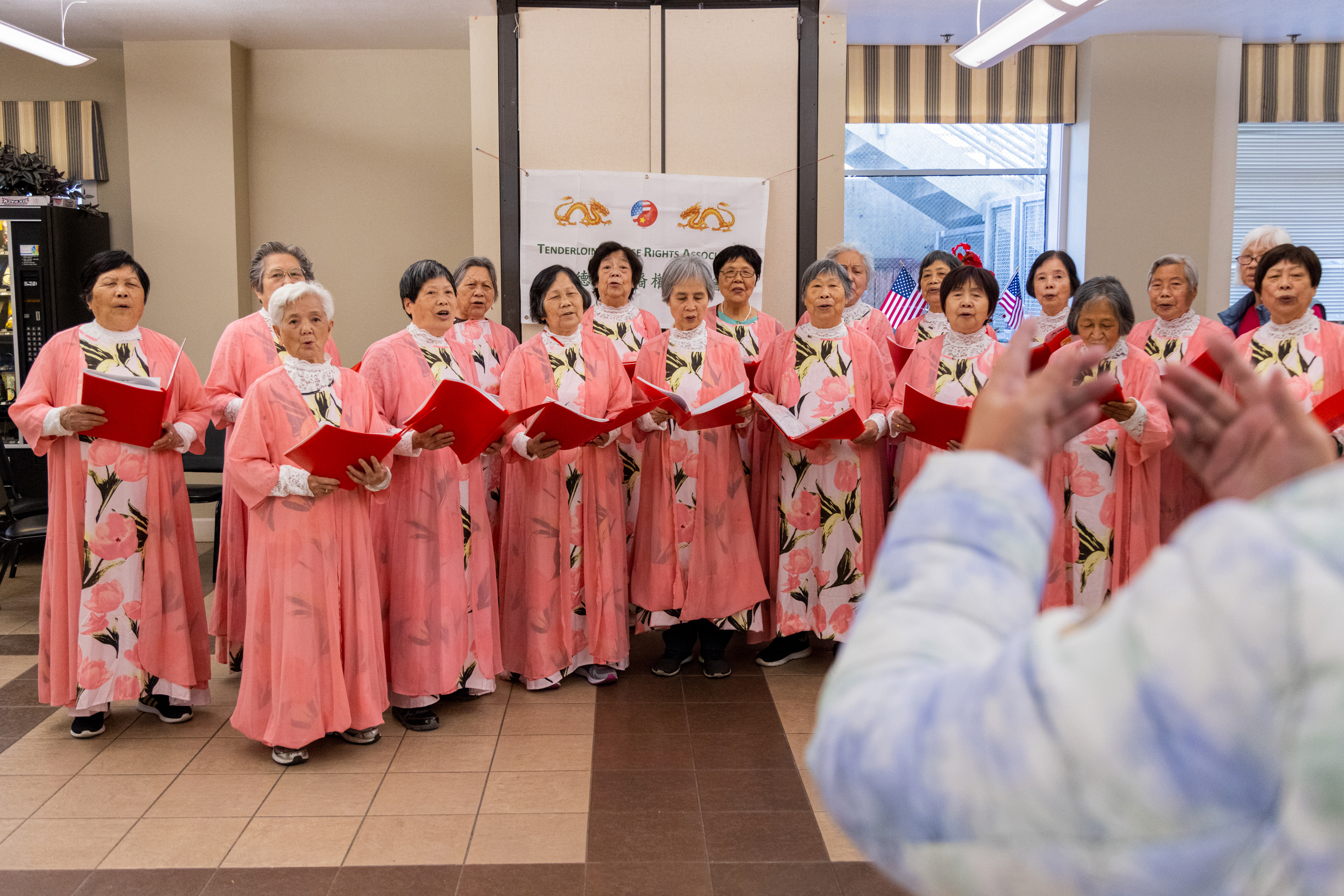 A choir of elderly women in pink floral robes sing passionately, holding red folders, conducted by a person with a raised hand, indoors.