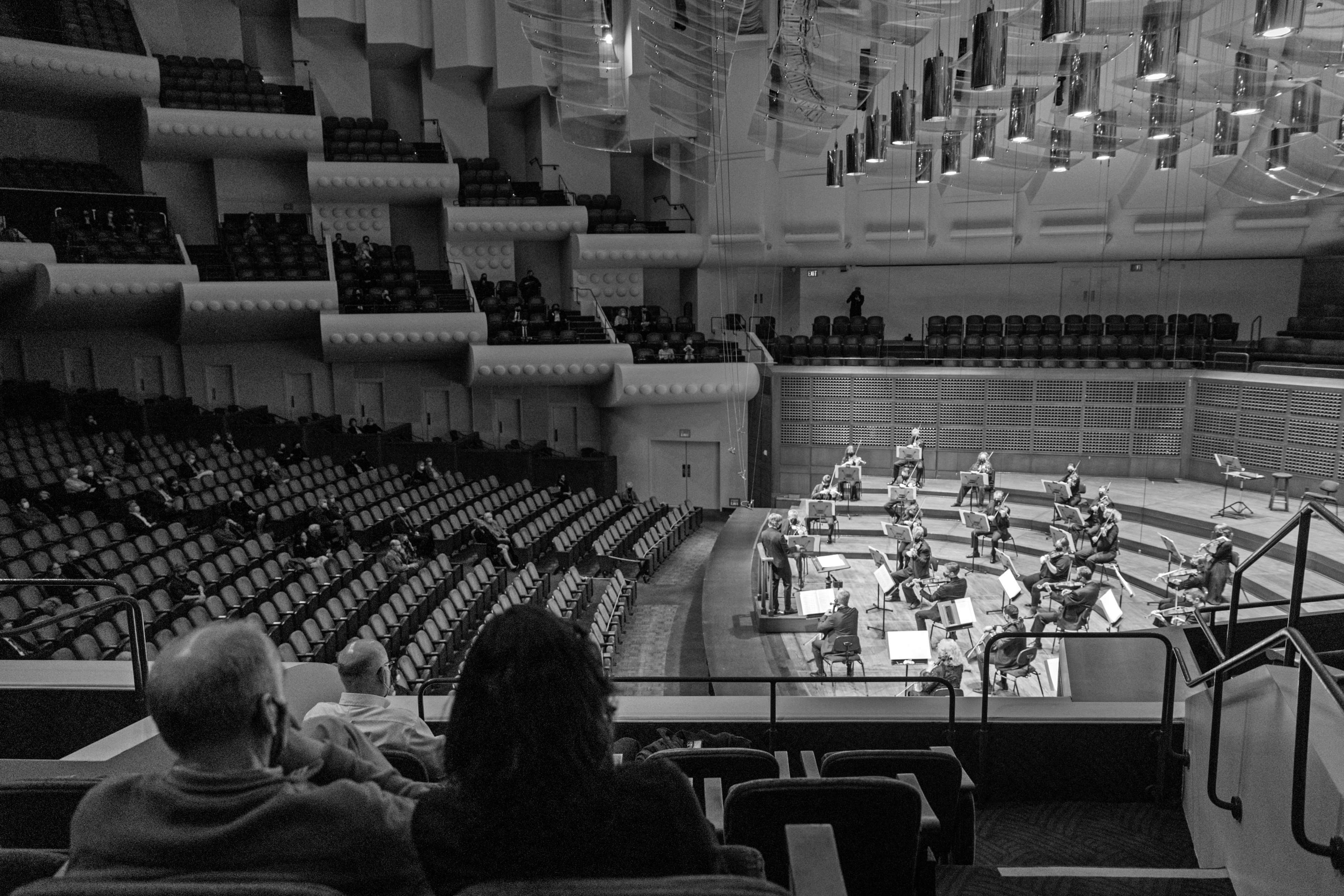 A black-and-white photo of a few audience members watching an orchestra play in a large, nearly empty modern concert hall.