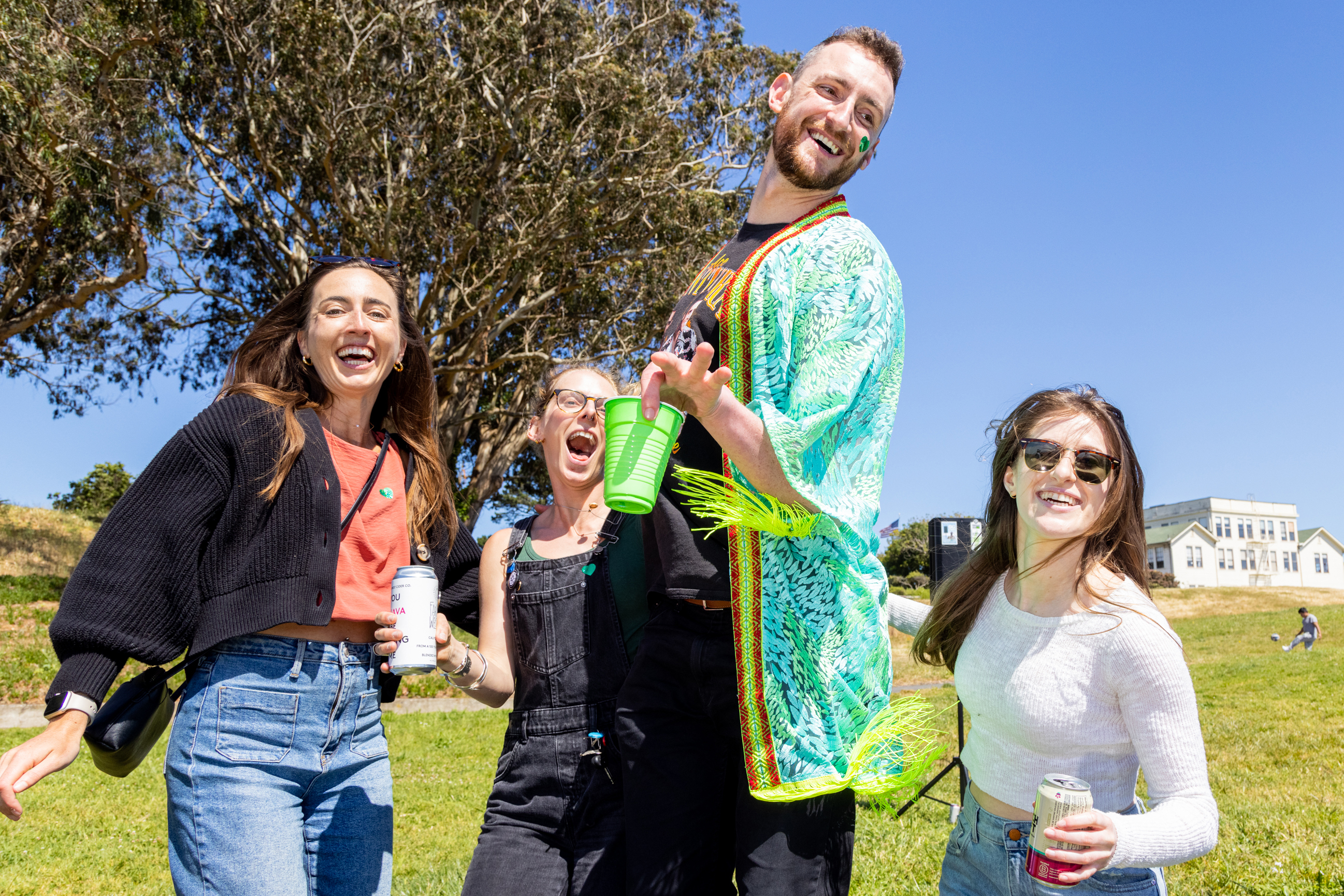 Four partygoers are laughing and enjoying a sunny day in a park, with drinks in their hands and a backdrop of lush greenery and a distant building.