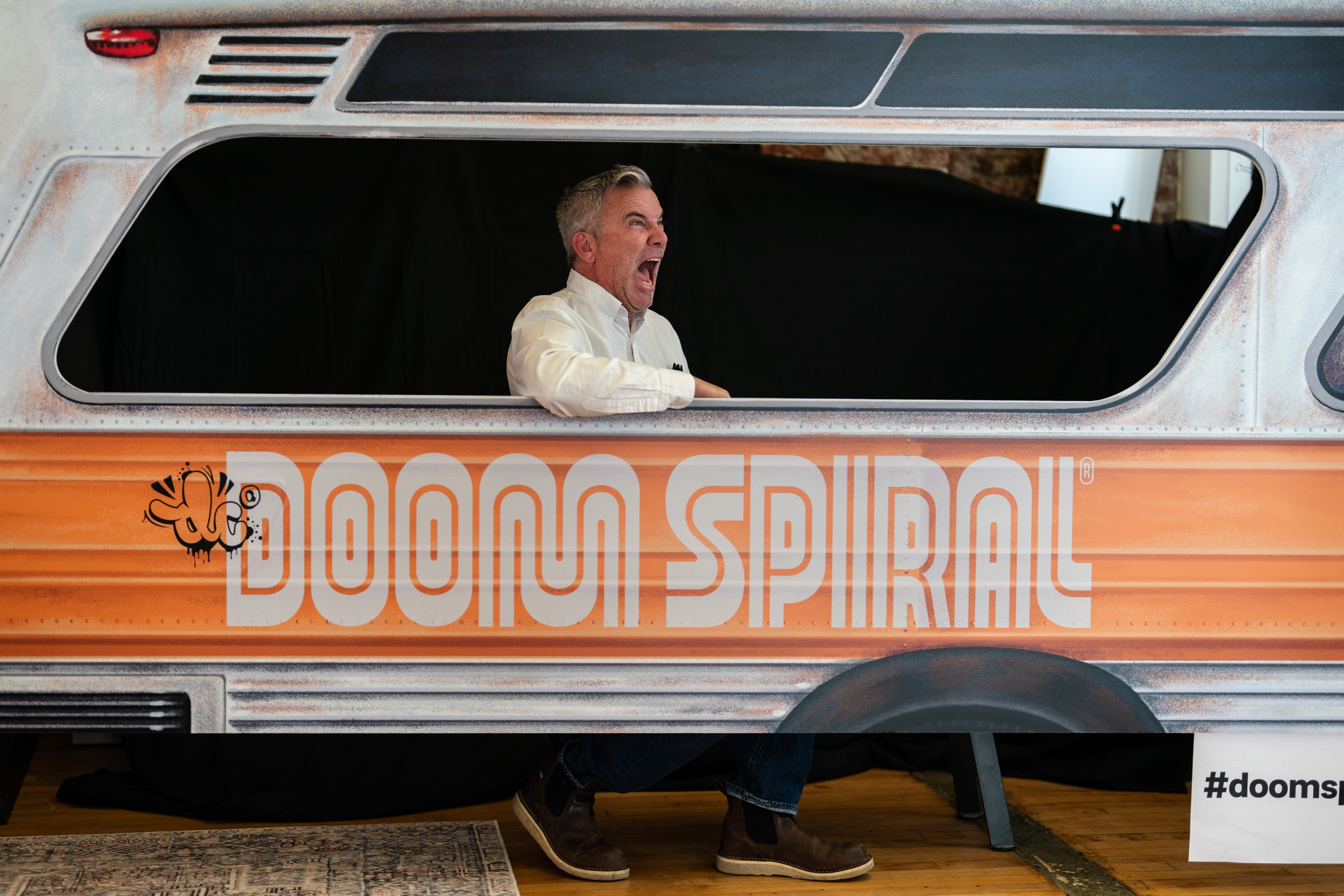A man joyfully leans out of a faux vehicle window with the words "DOOM SPIRAL" on it. The scene has a playful, animated atmosphere with a hashtag nearby.