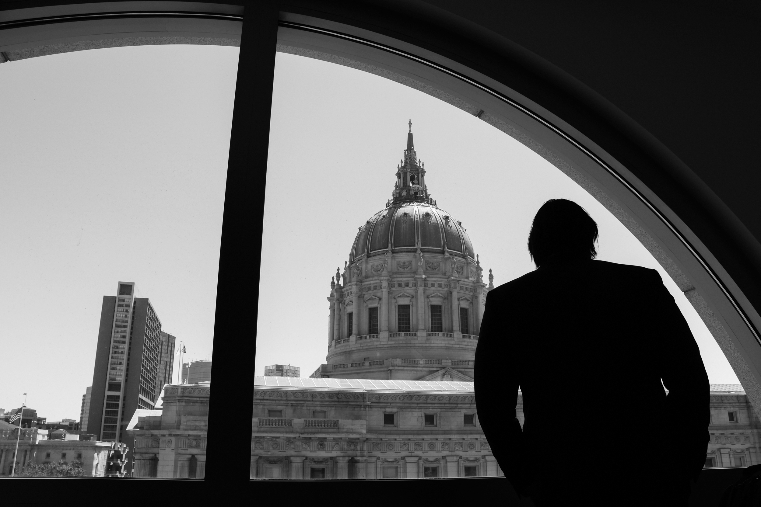 A silhouette of a man stands in front of a large window with an arched top, looking out at a domed building and surrounding cityscape in broad daylight.