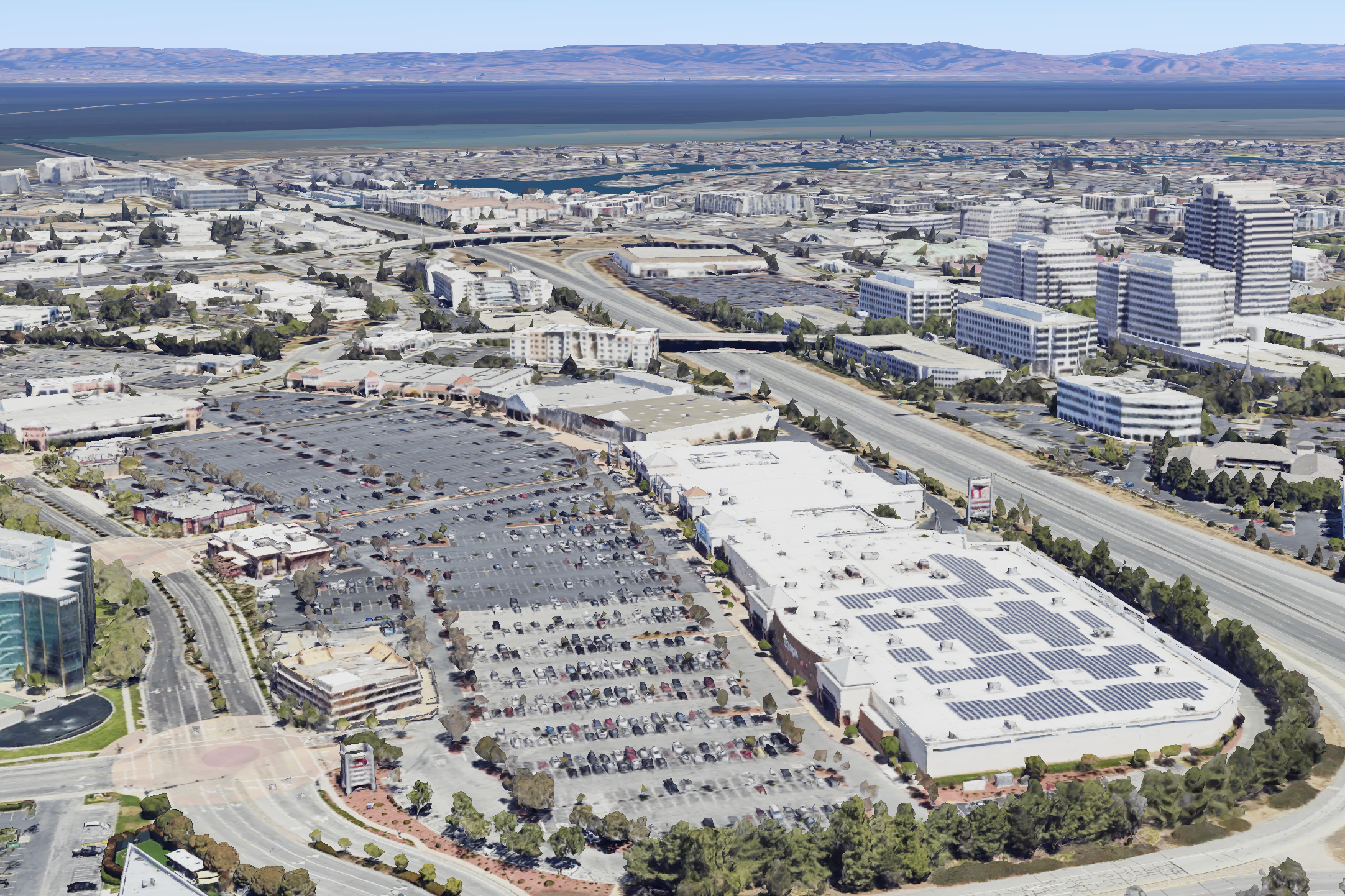The image shows an expansive urban area with large buildings, parking lots filled with cars, and highways. In the background, there is a view of a wide bay and distant mountains.