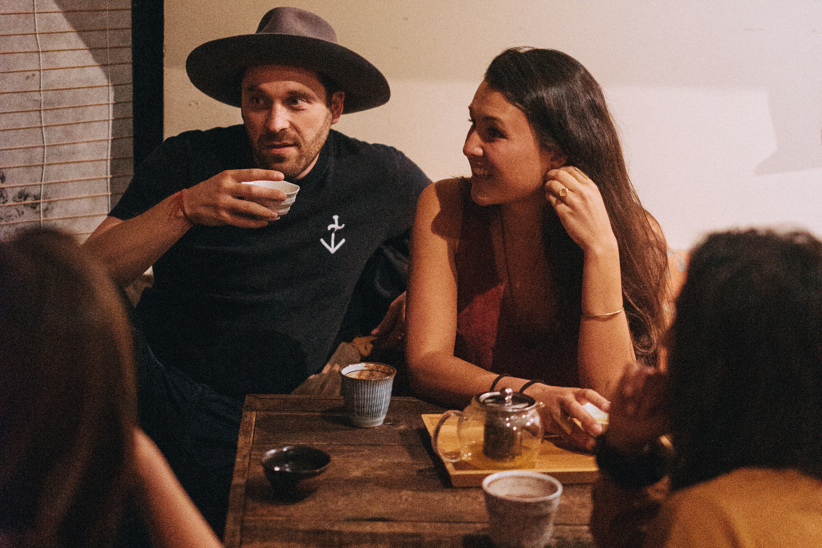 A man wearing a hat and a woman are sitting at a wooden table, chatting and enjoying drinks with others. The atmosphere is cozy and dimly lit.
