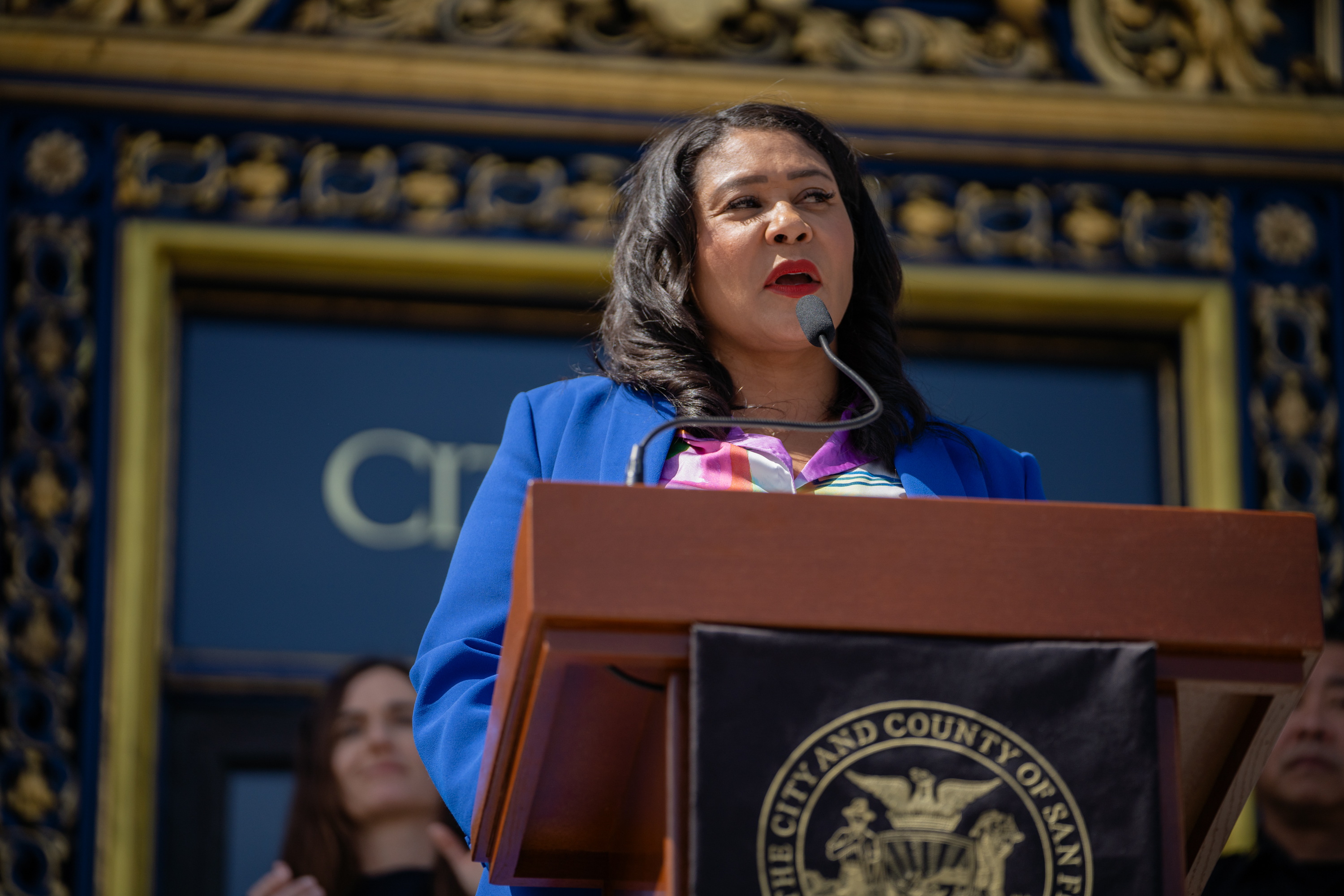 A woman in a blue blazer speaks at a podium with the City and County of San Francisco seal, with a decorative building and others in the background.