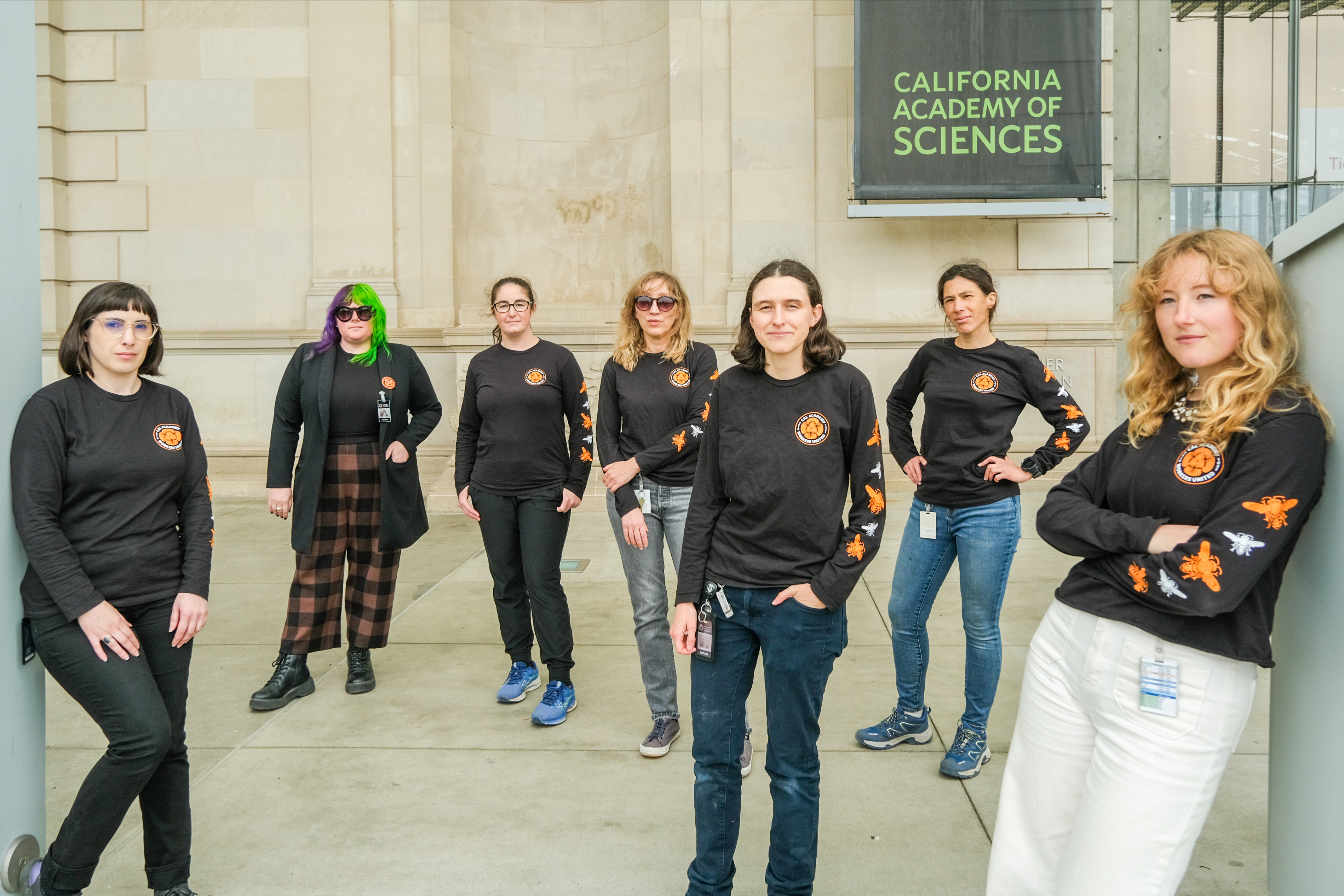Seven people in black shirts with orange logos stand confidently in front of the California Academy of Sciences.