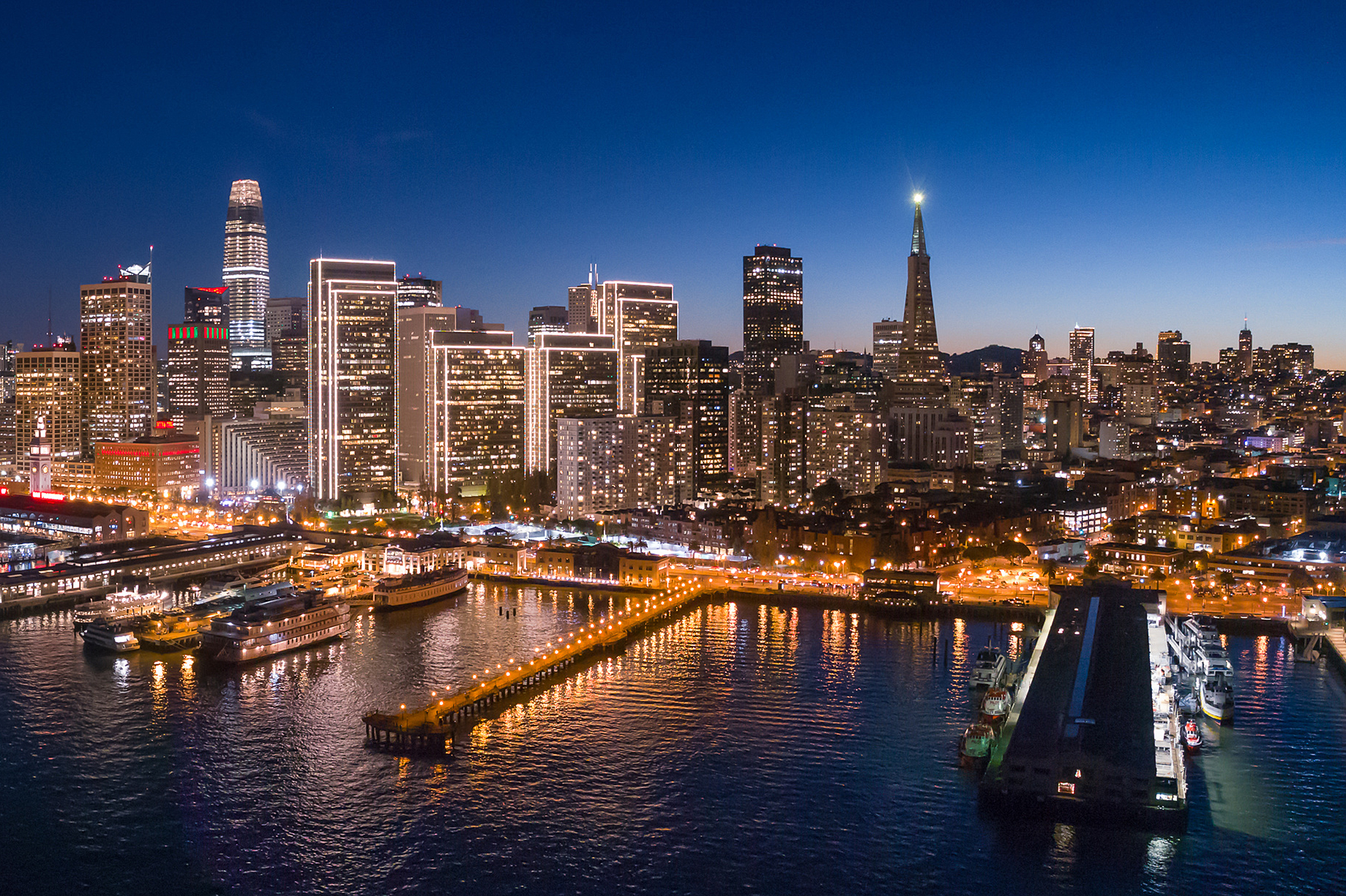 The image shows a vibrant city skyline at dusk with skyscrapers illuminated, reflecting in the water below. A pier with boats is visible in the foreground.