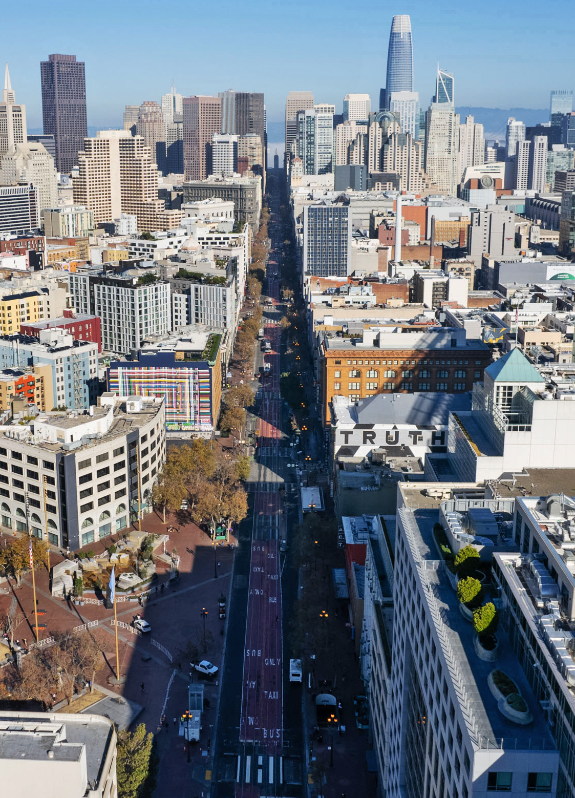 Aerial view of a tree-lined city street leading toward a dense cluster of downtown skyscrapers, with a building marked "TRUTH" on the right.