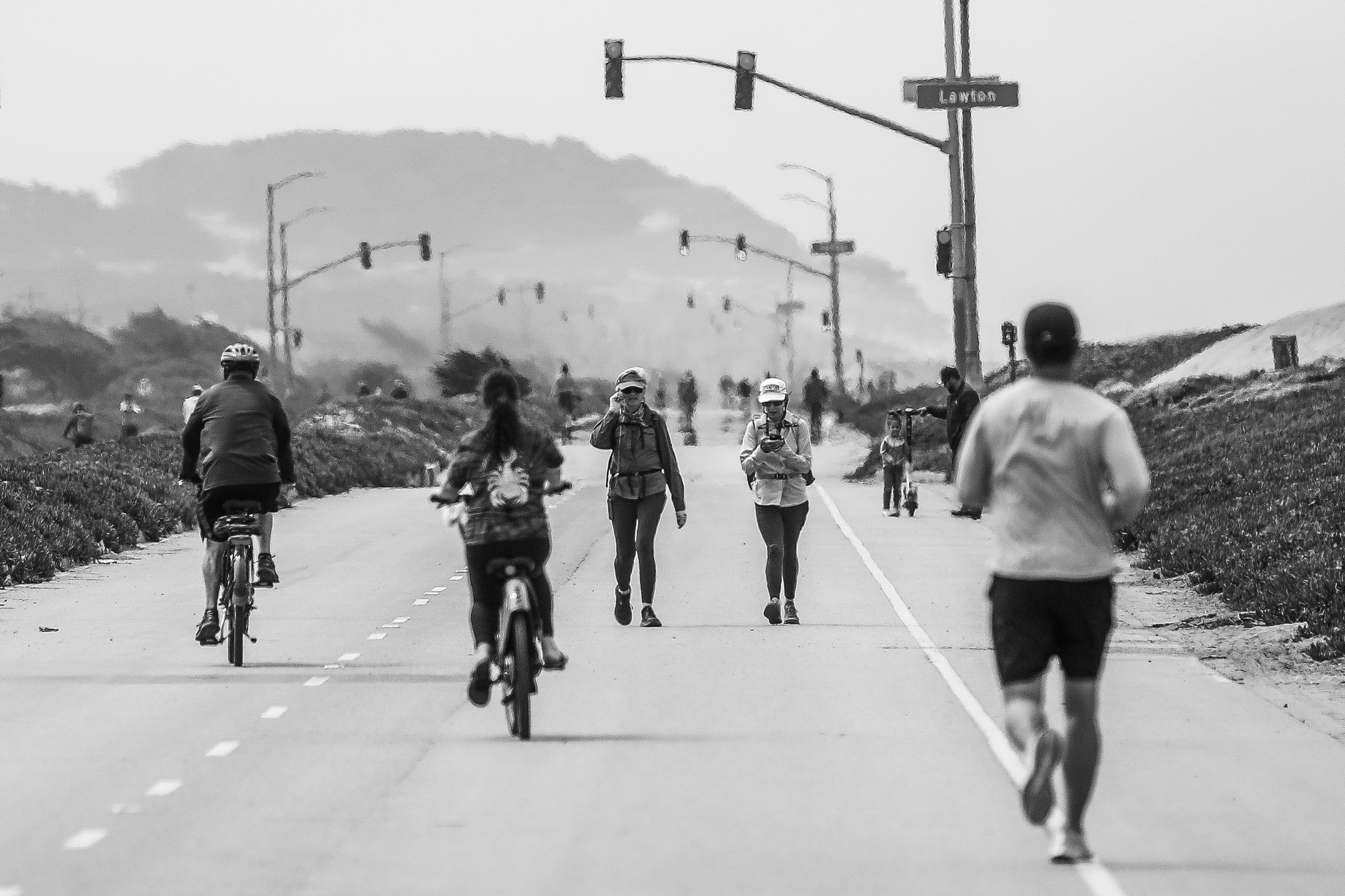 The image shows a grayscale scene of a shared road with cyclists and pedestrians, some jogging or walking dogs. Traffic lights and distant hills are visible ahead.