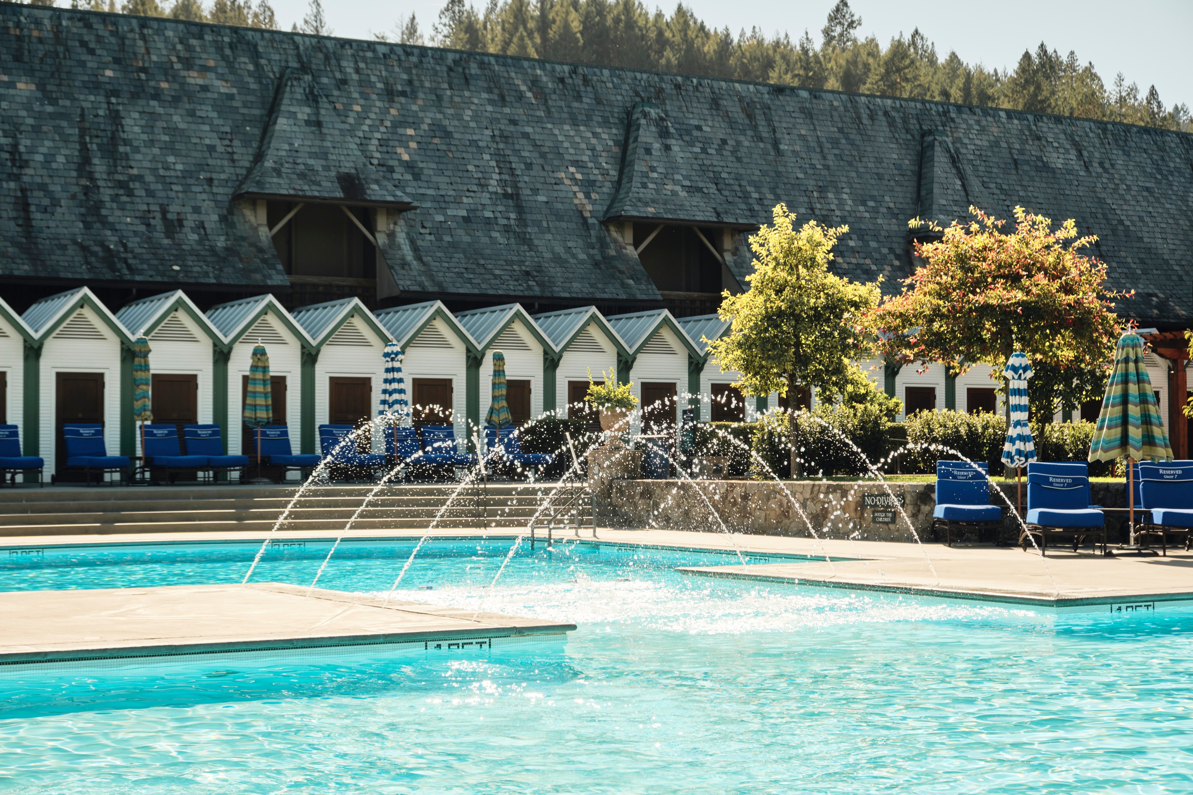 The image depicts a serene outdoor pool with fountains, surrounded by blue lounge chairs, parasols, and white beach-style cabins backed by a building with a shingled roof.