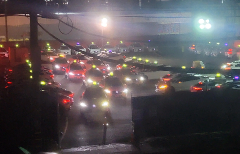 The image captures a congestion of cars at night, under a light drizzle, with glowing headlights and colorful brake lights reflecting off the wet ground.