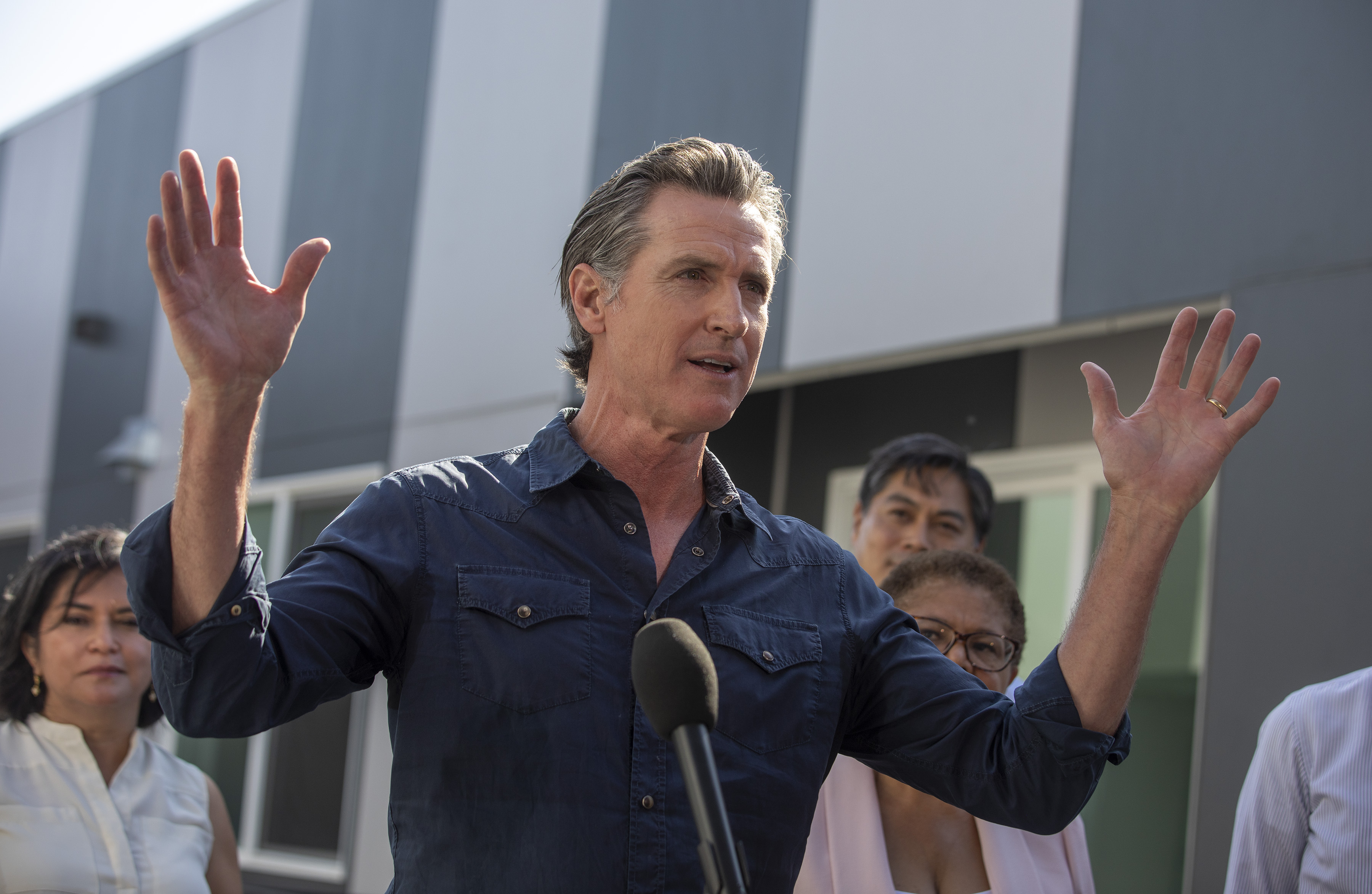 A man in a blue shirt is speaking outdoors, gesturing with both hands raised. He is surrounded by several people, and there's a building with large windows behind them.