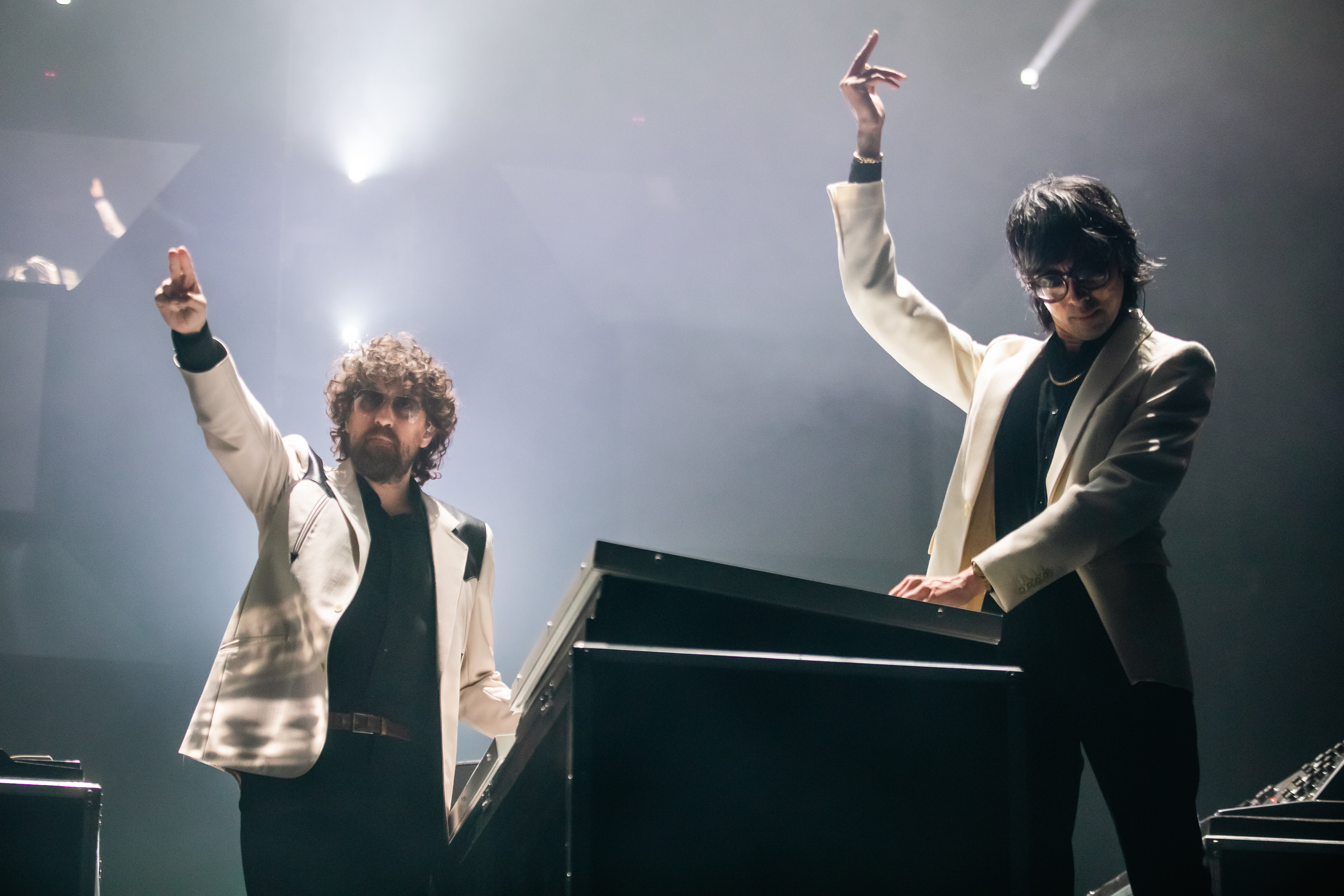 Two musicians in white jackets perform on stage, one pointing up, the other at a keyboard, amidst dramatic lighting.