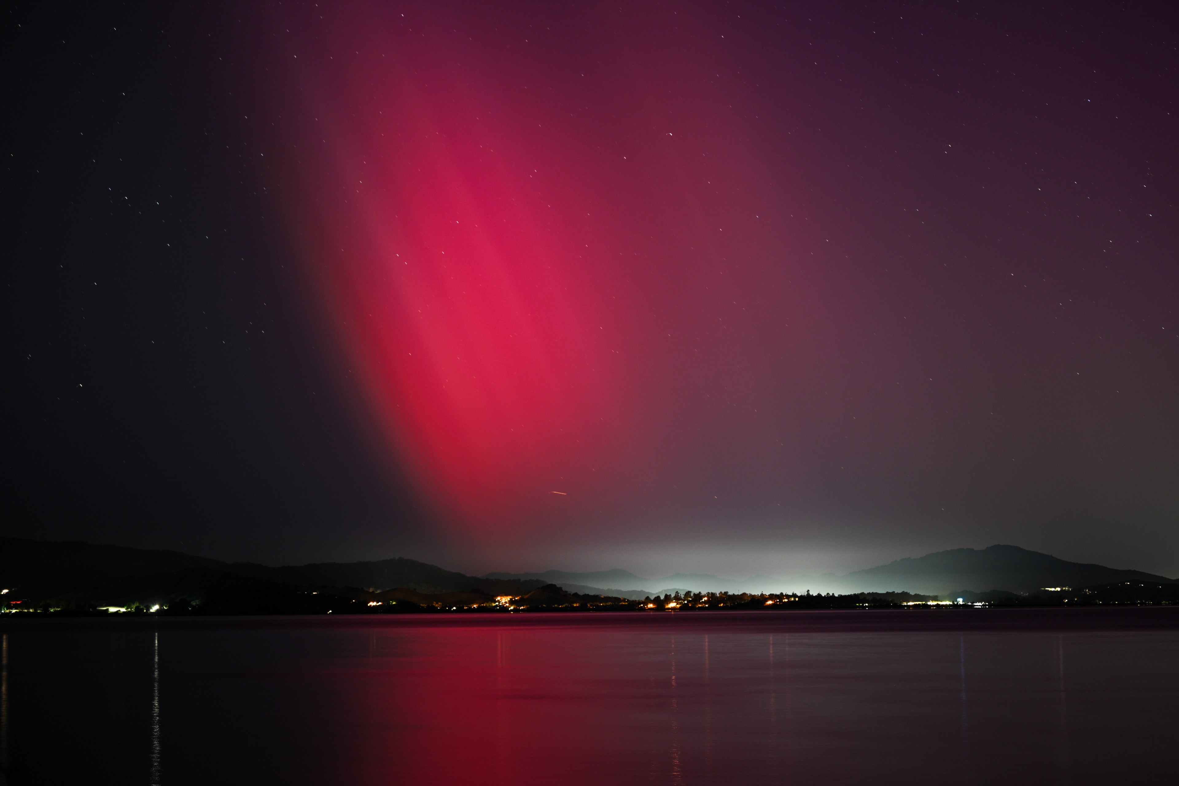 Red aurora-like lights streak across the night sky above glowing city lights and a calm lake.
