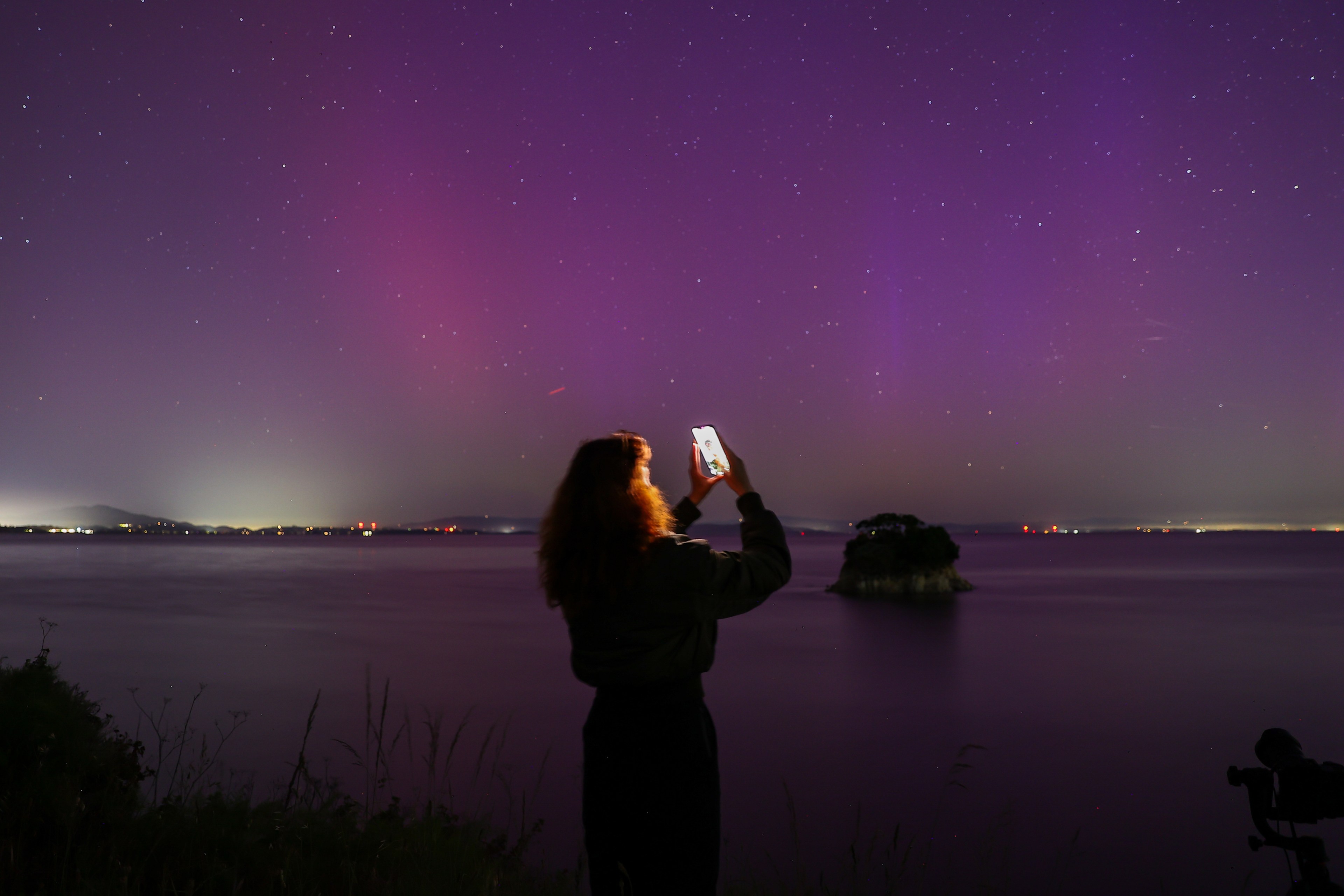 A person is taking a picture of aurora-like lights in a starry night sky over a calm lake with their phone.