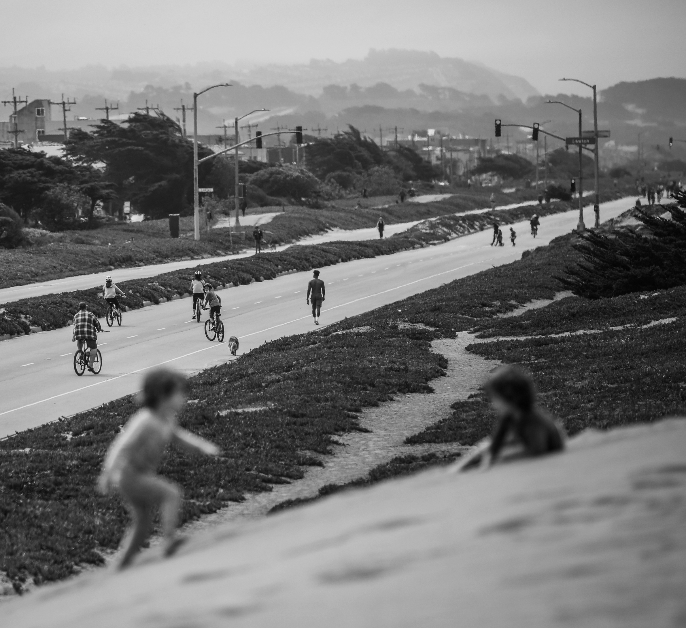 The black-and-white image depicts a street scene with cyclists and pedestrians along a hilly road. In the foreground, two children play on a grassy slope.