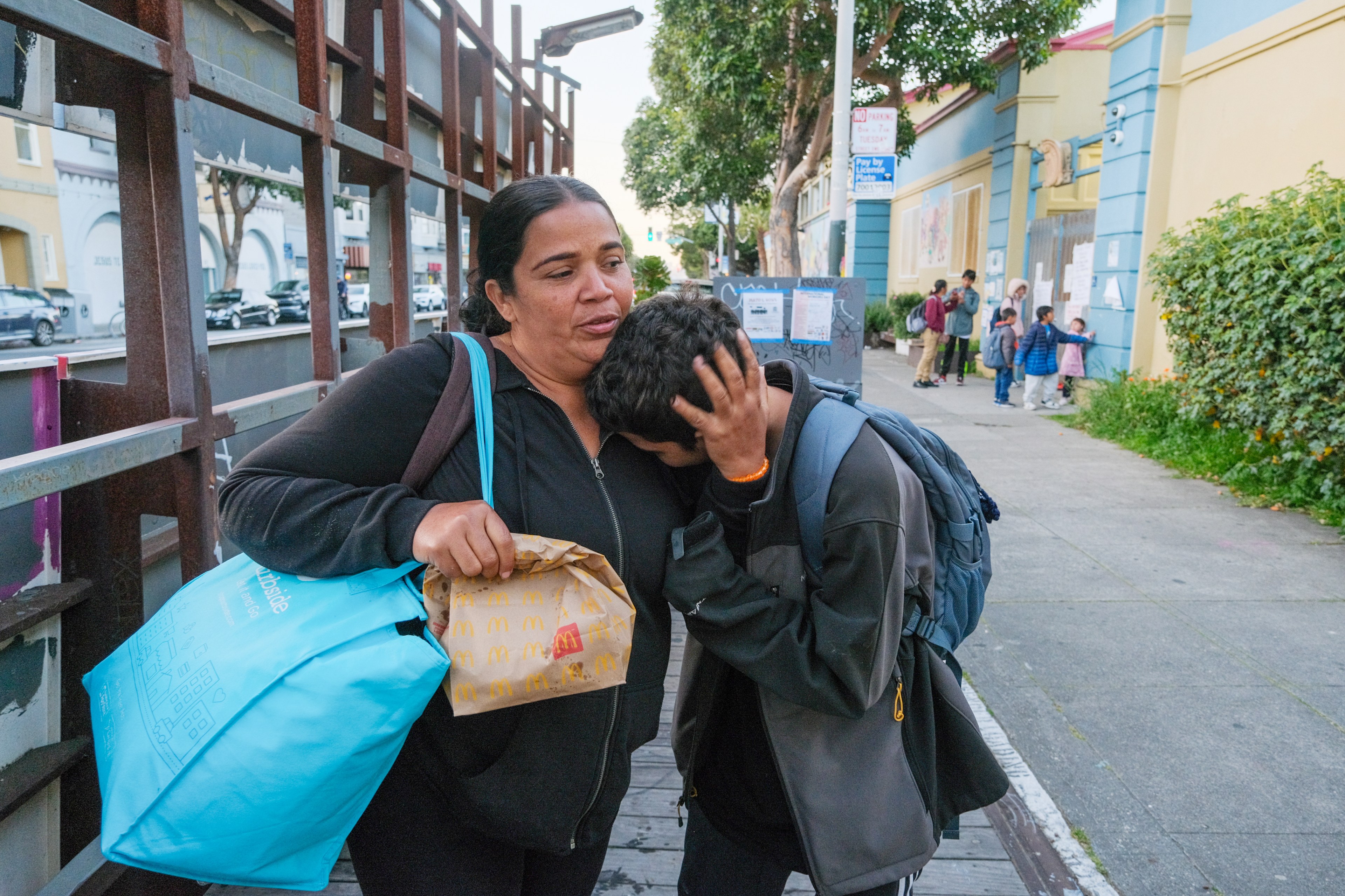 A woman comforts a young person on a sidewalk; both seem distressed, with a fast-food bag in view.