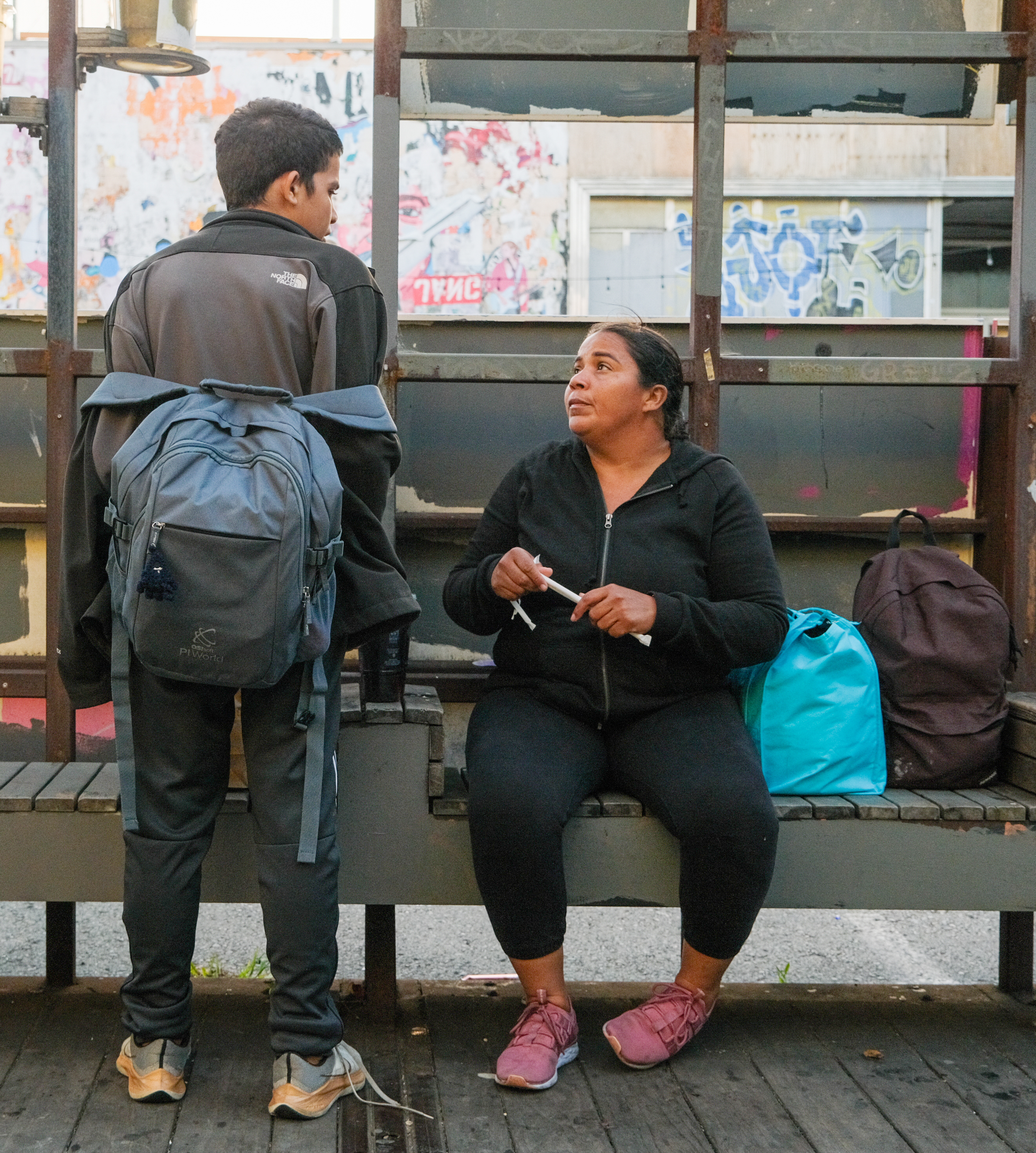 A boy with a backpack facing a seated woman with headphones, both engaging in conversation at a graffiti-lined train station.