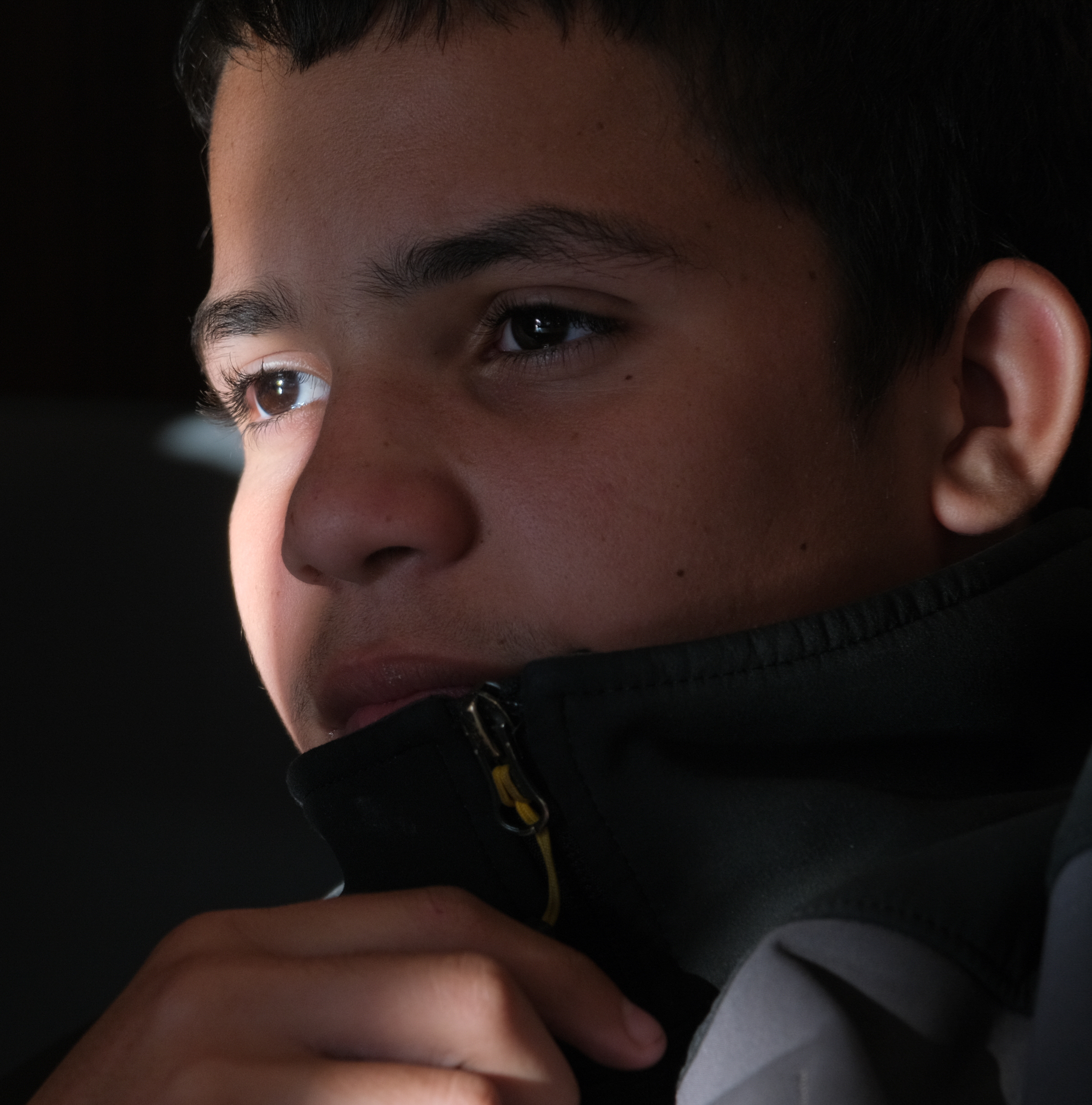 A close-up of a pensive young boy with his chin resting on his hand, wearing a zipped-up jacket.
