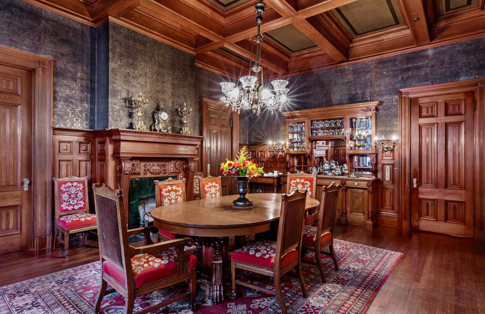 An ornate dining room with a round wooden table, chairs, a bar area, a fireplace, and rich wood paneling.
